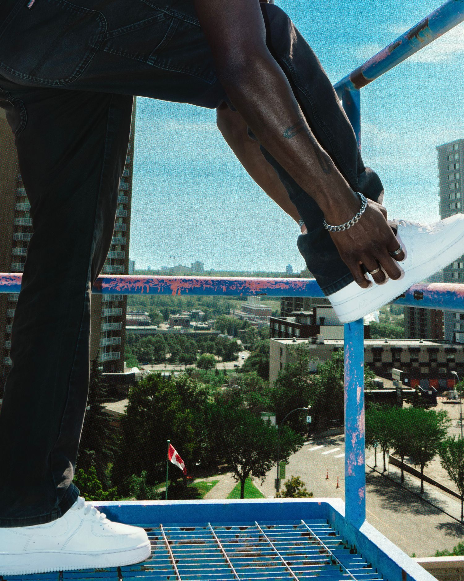 Close-up of a person in black jeans and white sneakers sitting on a blue railing above a city.
