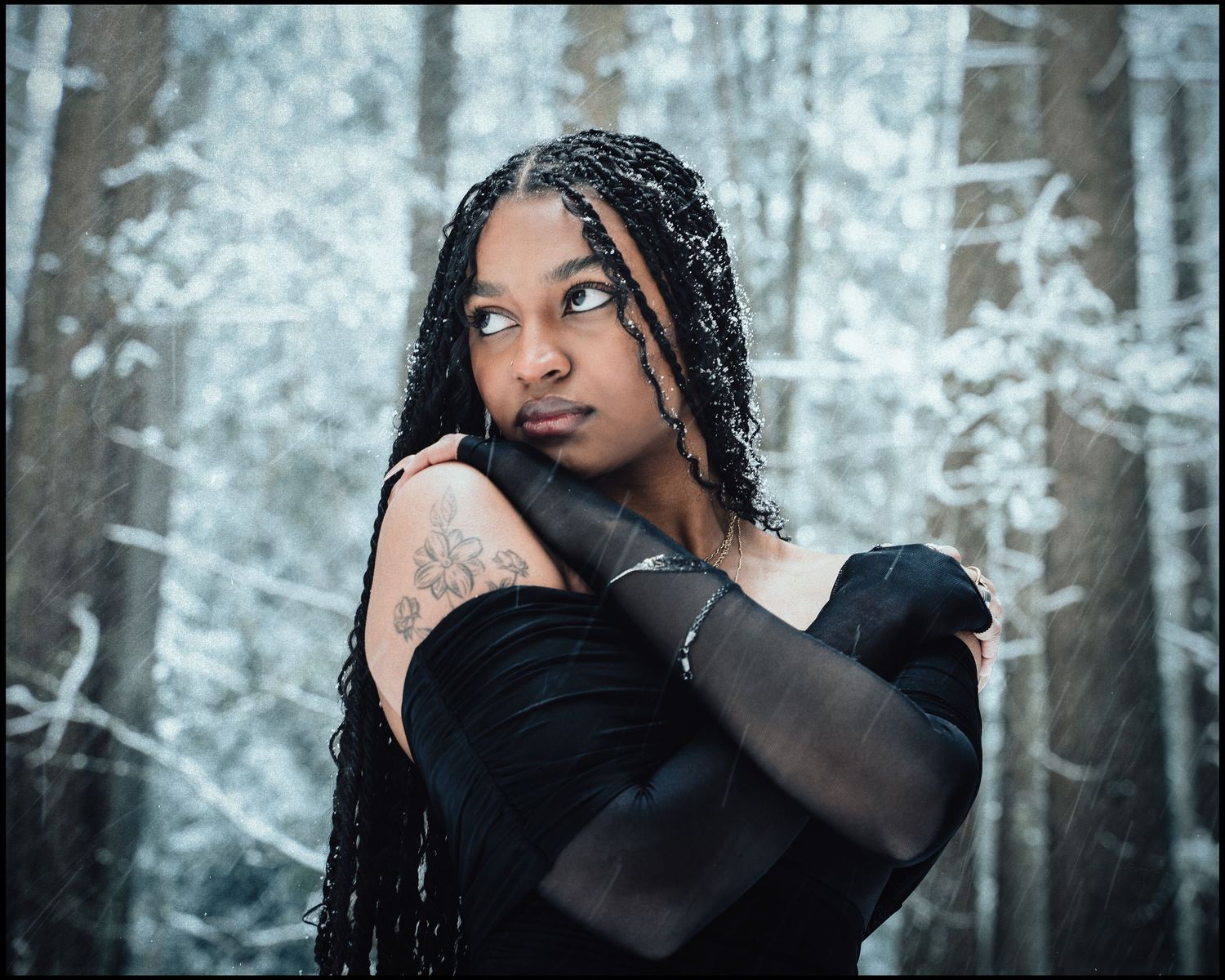 Young woman with braids and tattoo posing in snowy forest wearing black gloves.
