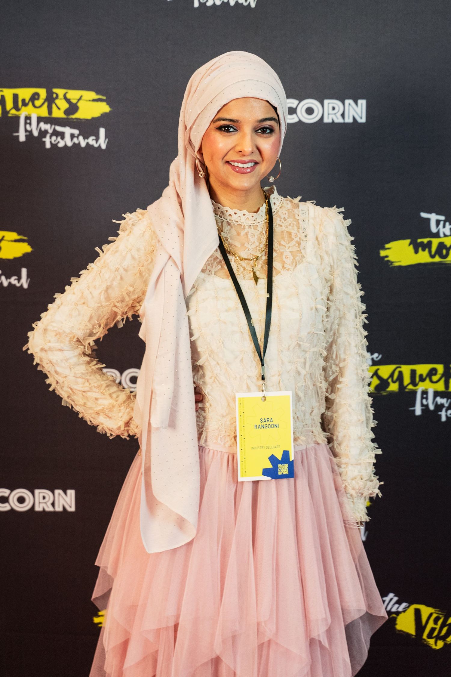 Young woman in pink hijab and white feathered jacket poses at the Mosque Film Festival red carpet.