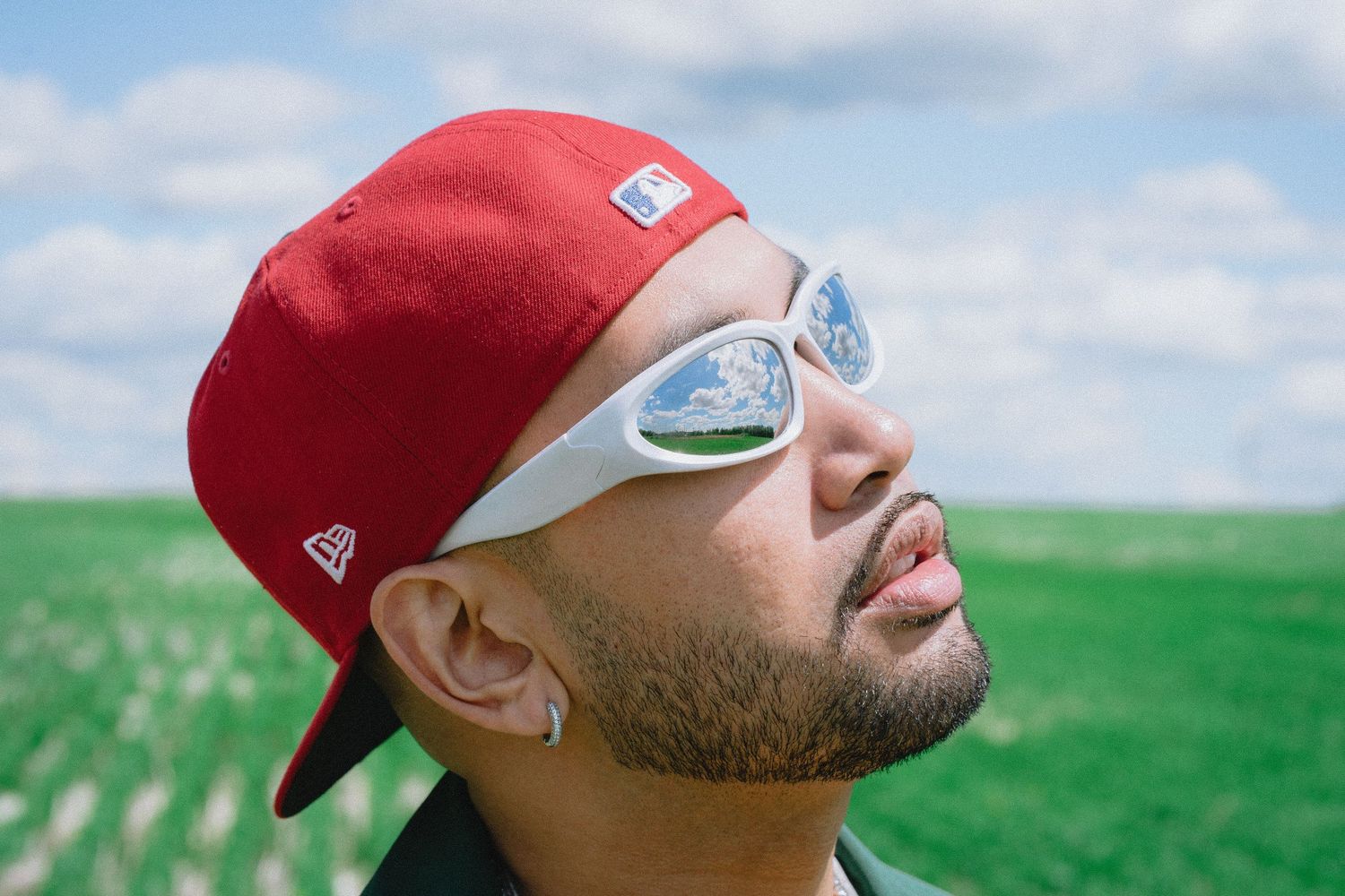 Close-up of man wearing red New Era cap and futuristic silver sunglasses looking up at cloudy sky.