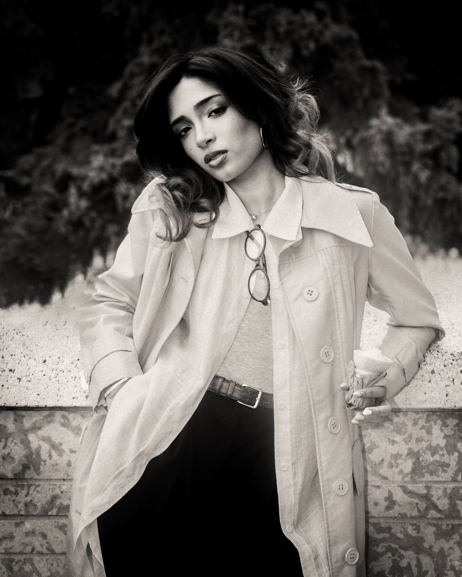Elegant woman in white trench coat and black pants poses confidently in dramatic black and white portrait.