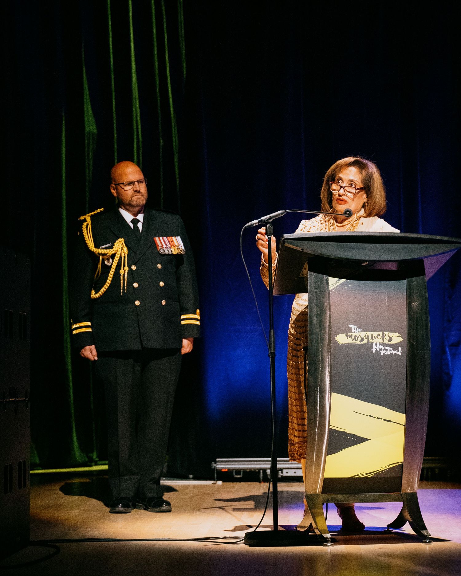Military officer in dress uniform stands beside a woman speaking at a podium during a formal ceremony on stage.