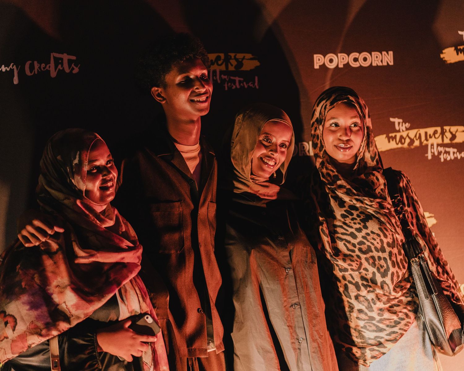 Four people wearing hijabs pose together smiling in front of a Popcorn festival backdrop under warm lighting.