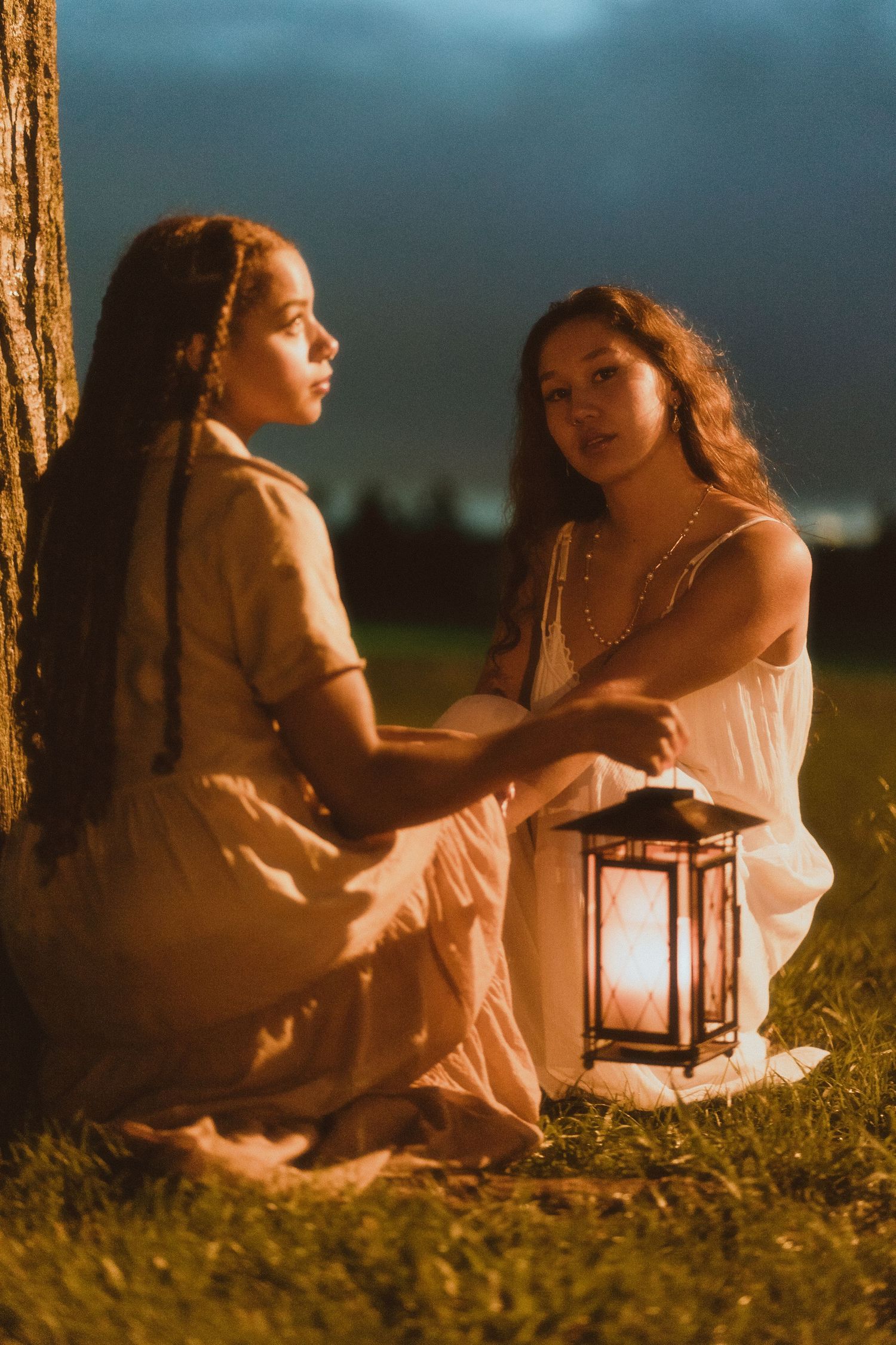 Two women in flowing dresses share a quiet moment by a glowing lantern under a tree at night.