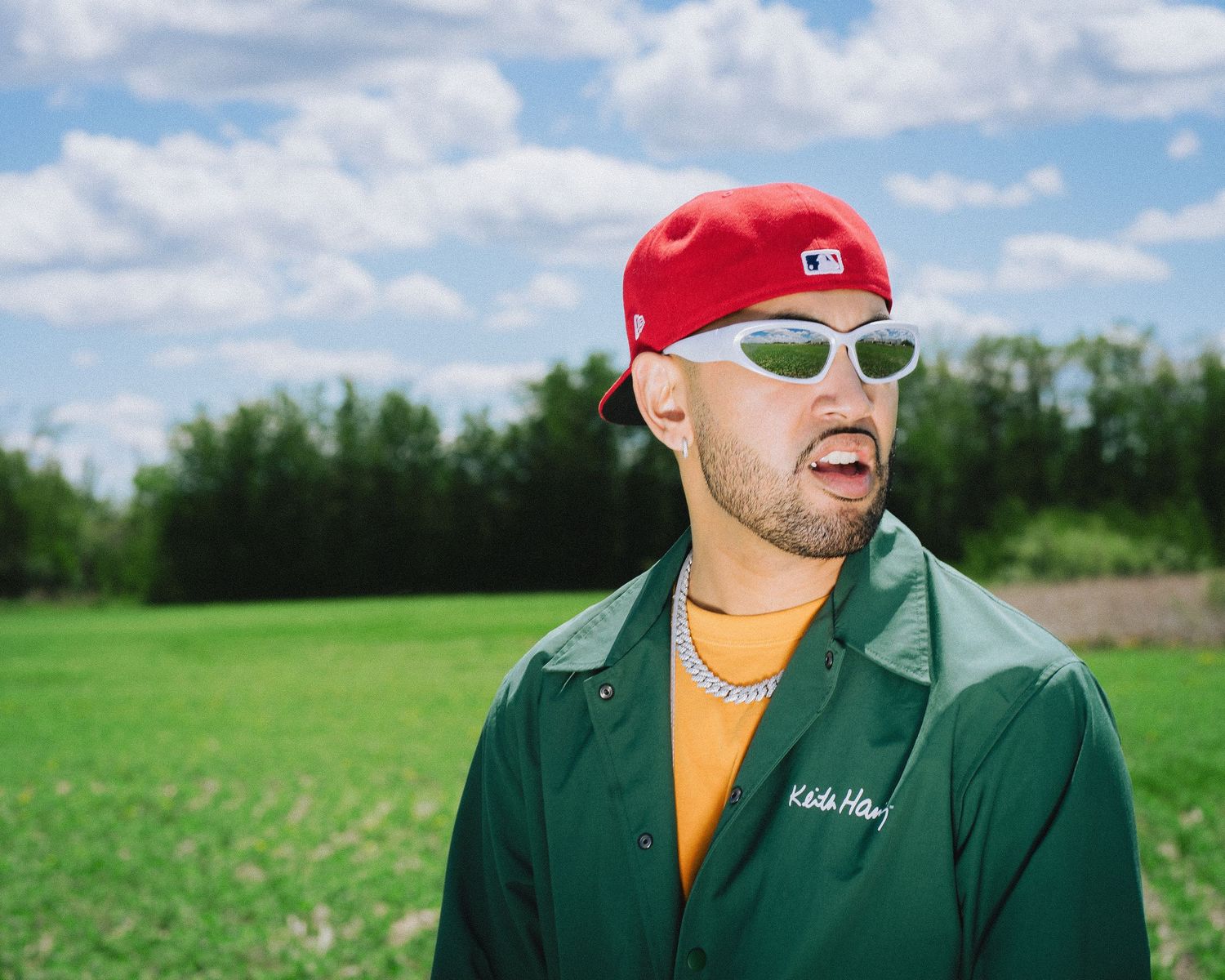 Man in red baseball cap, green jacket, and white sunglasses laughing outdoors in a green field.