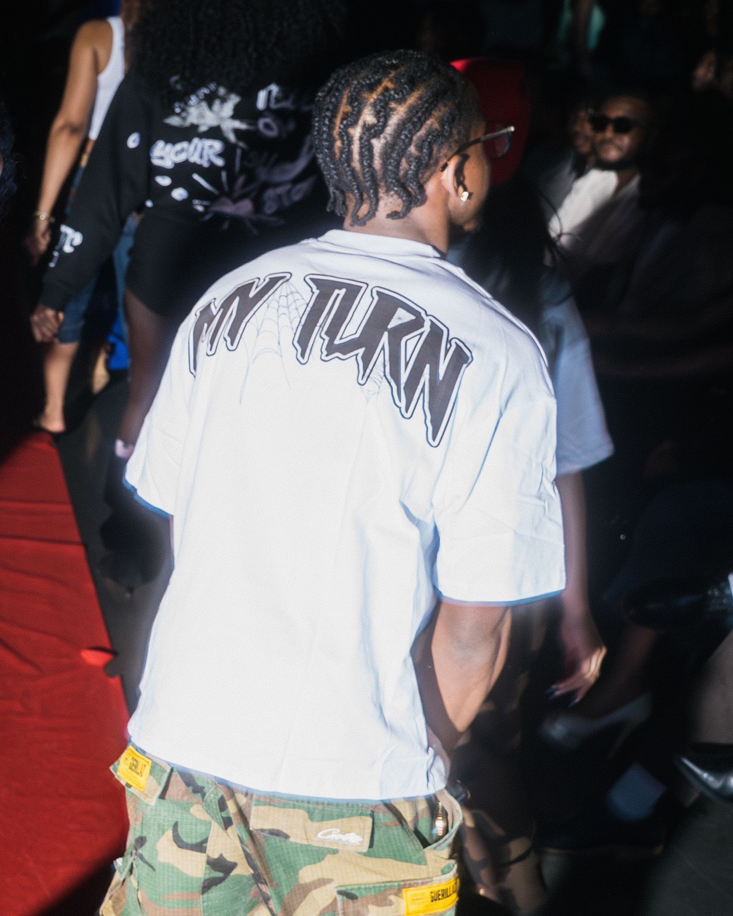 Male attendee wears white 'My Turn' graphic tee with camo pants at urban streetwear fashion show.