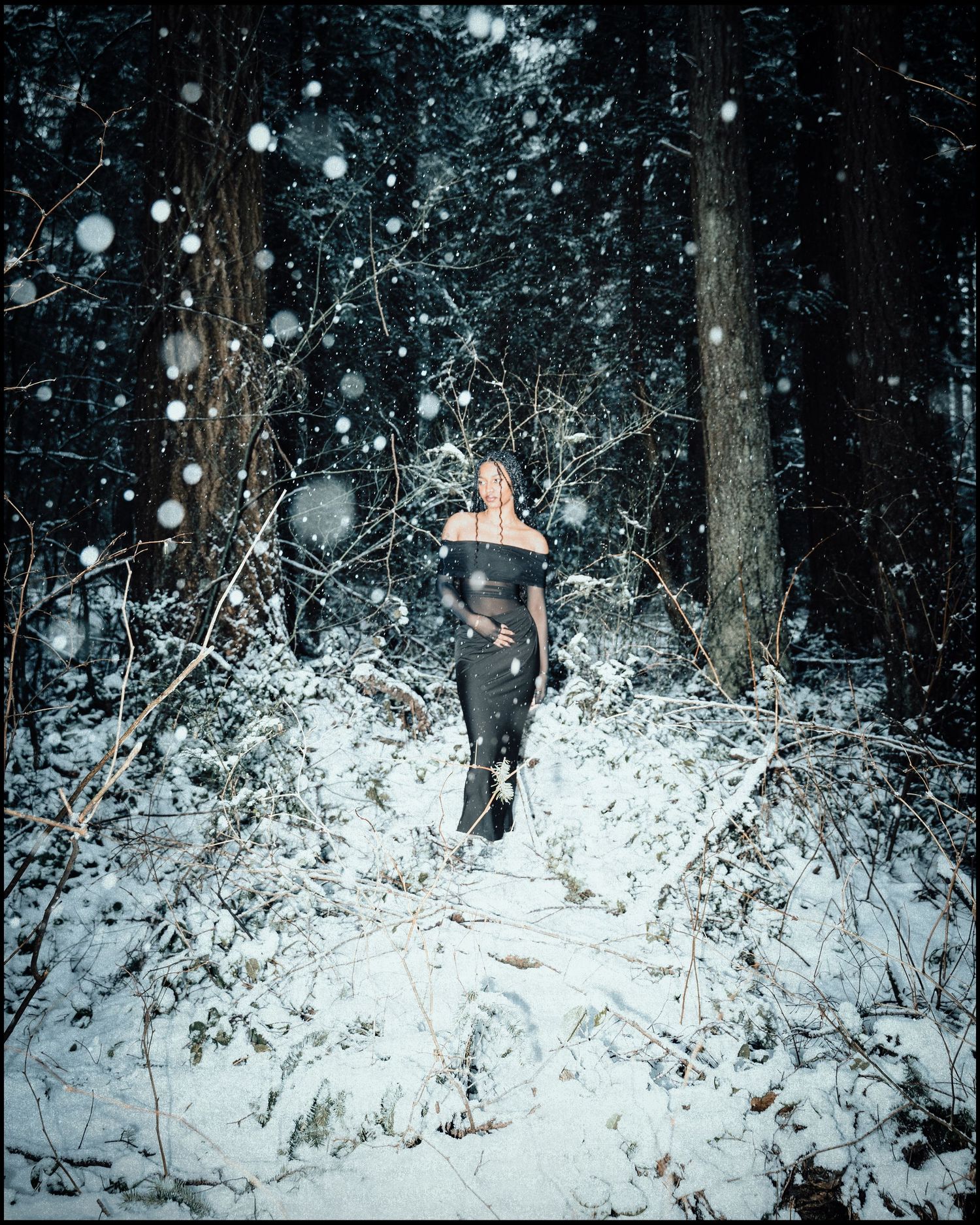 Woman in black off-shoulder outfit walking through snow-covered forest path.