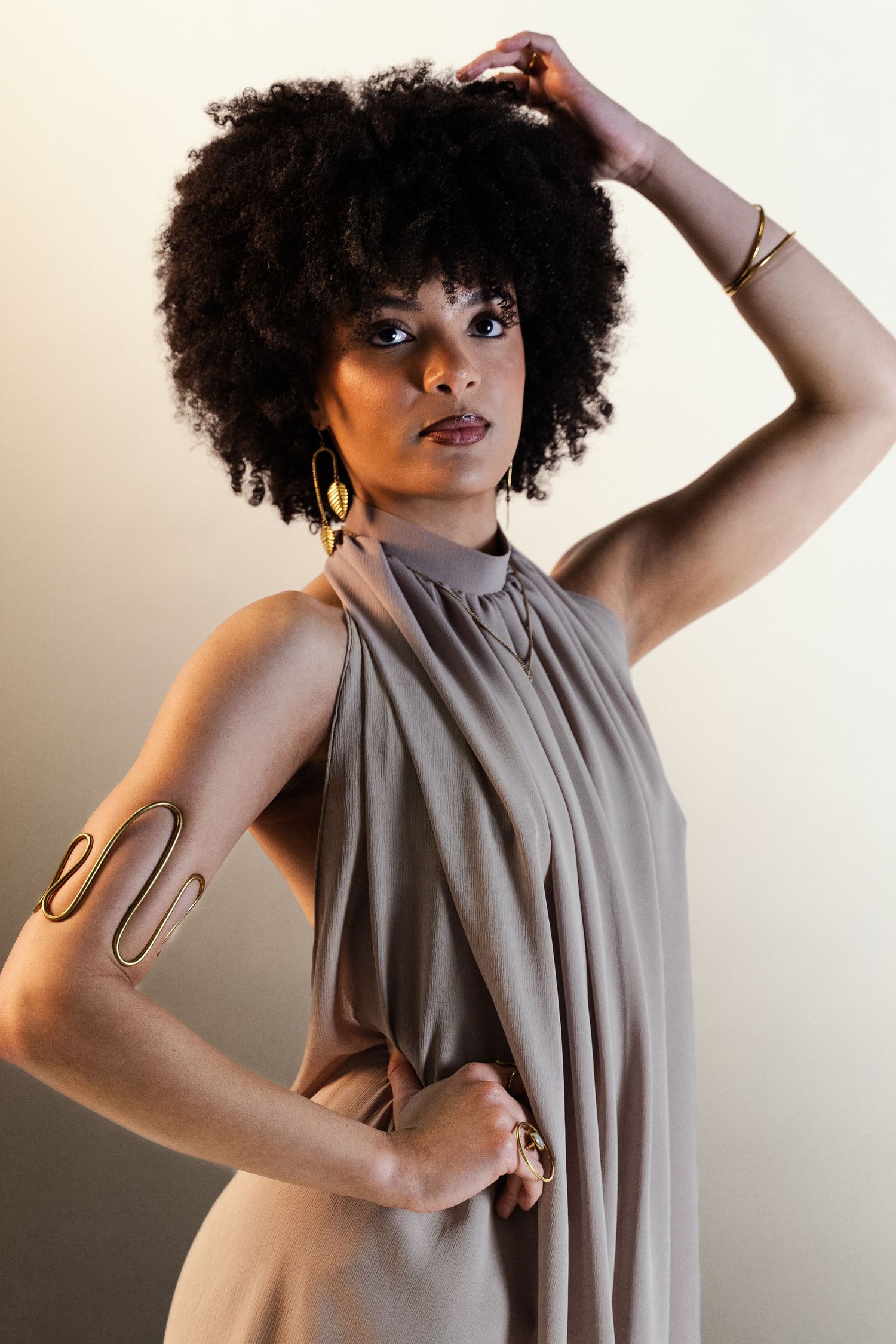 Model in taupe pleated halter dress poses with hand in afro hair, wearing gold hoop earrings and arm cuff.