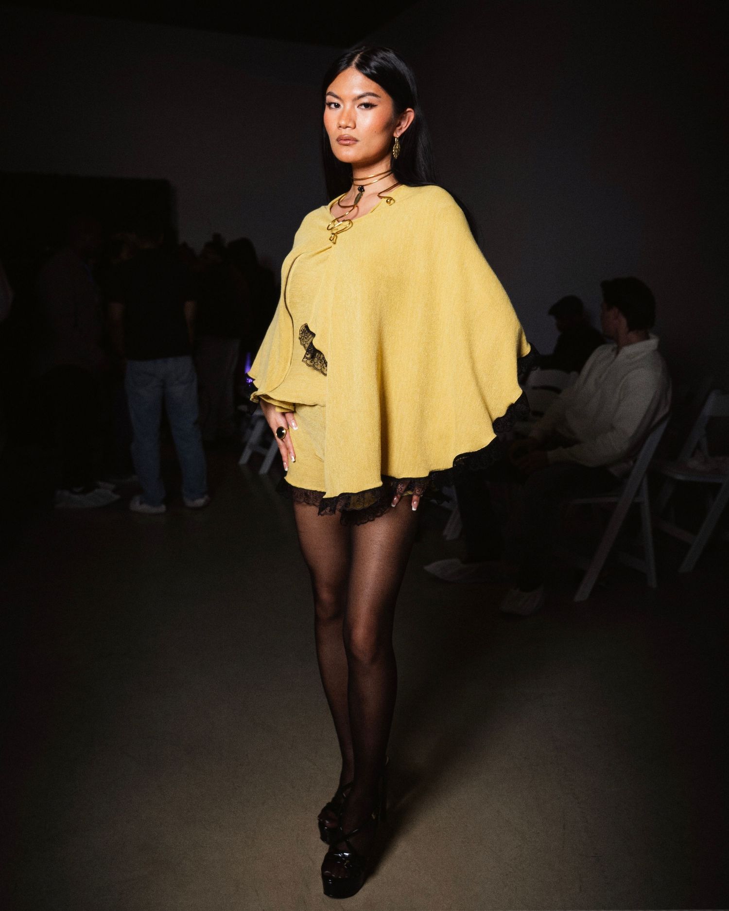 Model wearing yellow cape top with lace-up neckline, black shorts and tights at fashion show.
