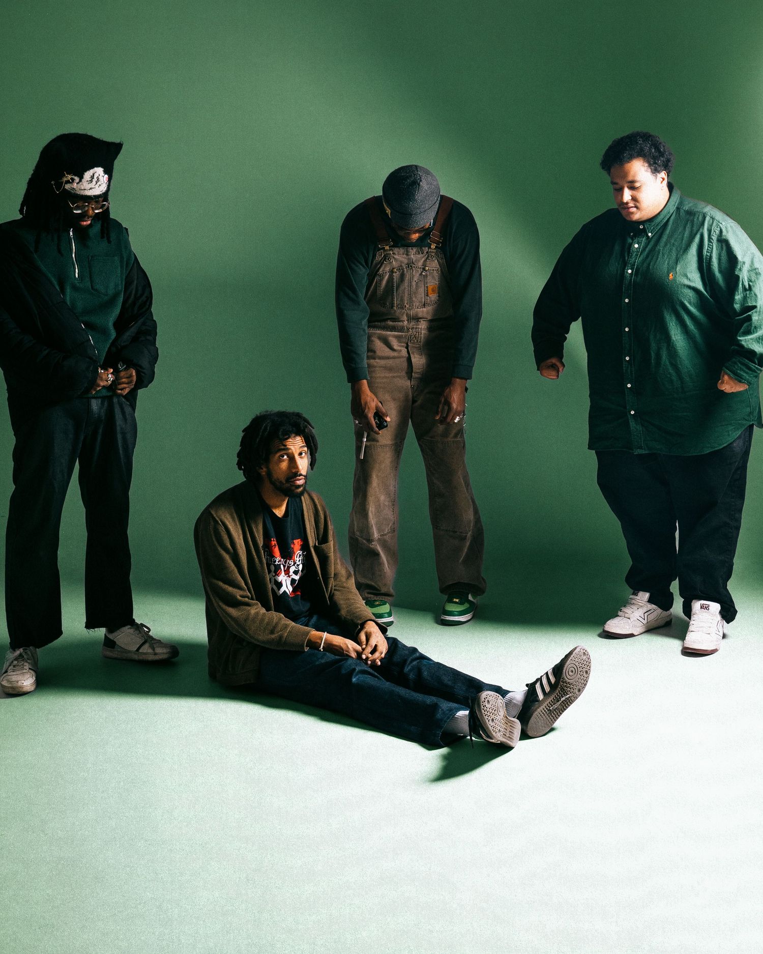 Four men posing in a green studio, one seated on the floor and three standing in casual streetwear.