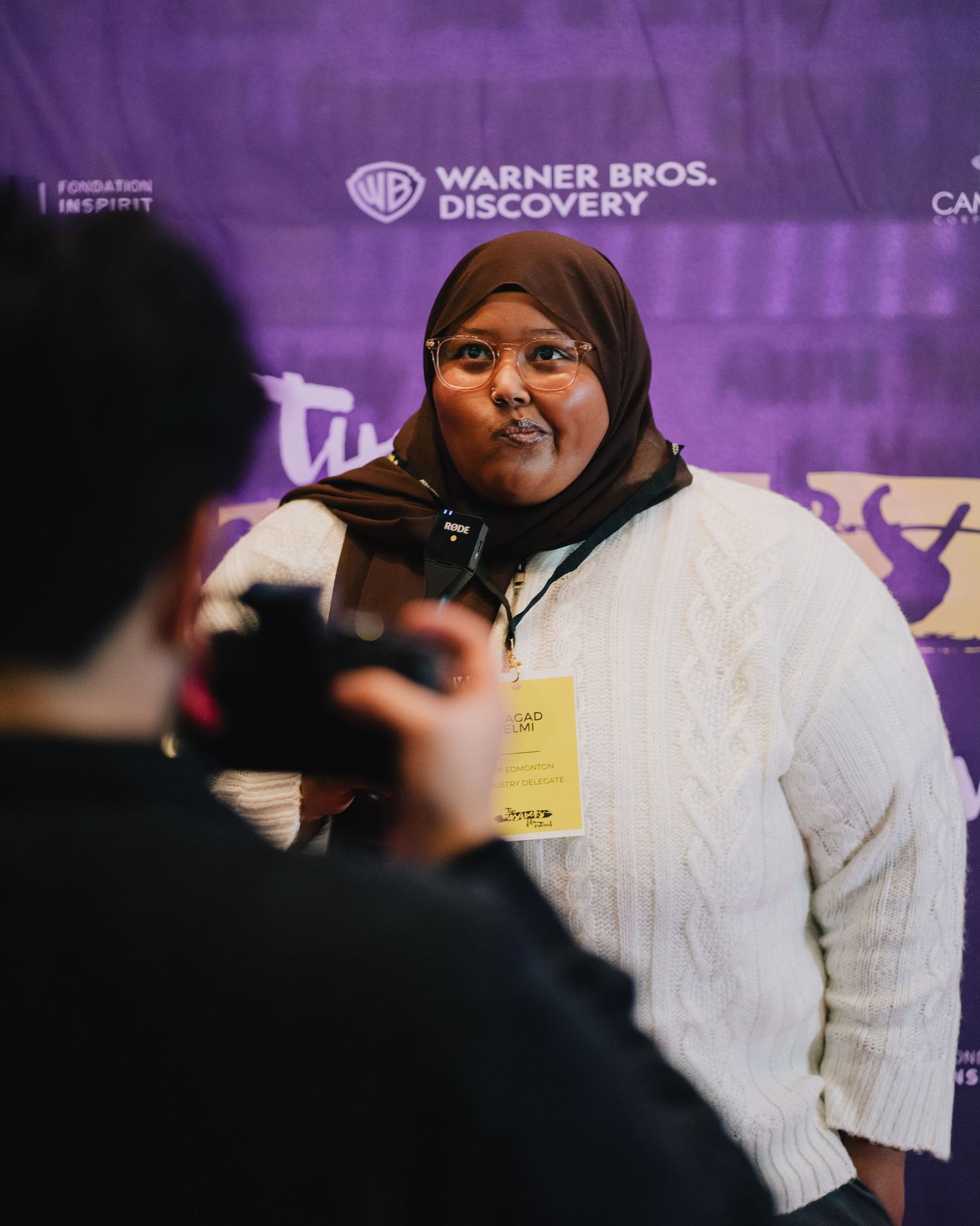 Woman wearing hijab and yellow lanyard poses for a photo in front of a Warner Bros. Discovery banner.