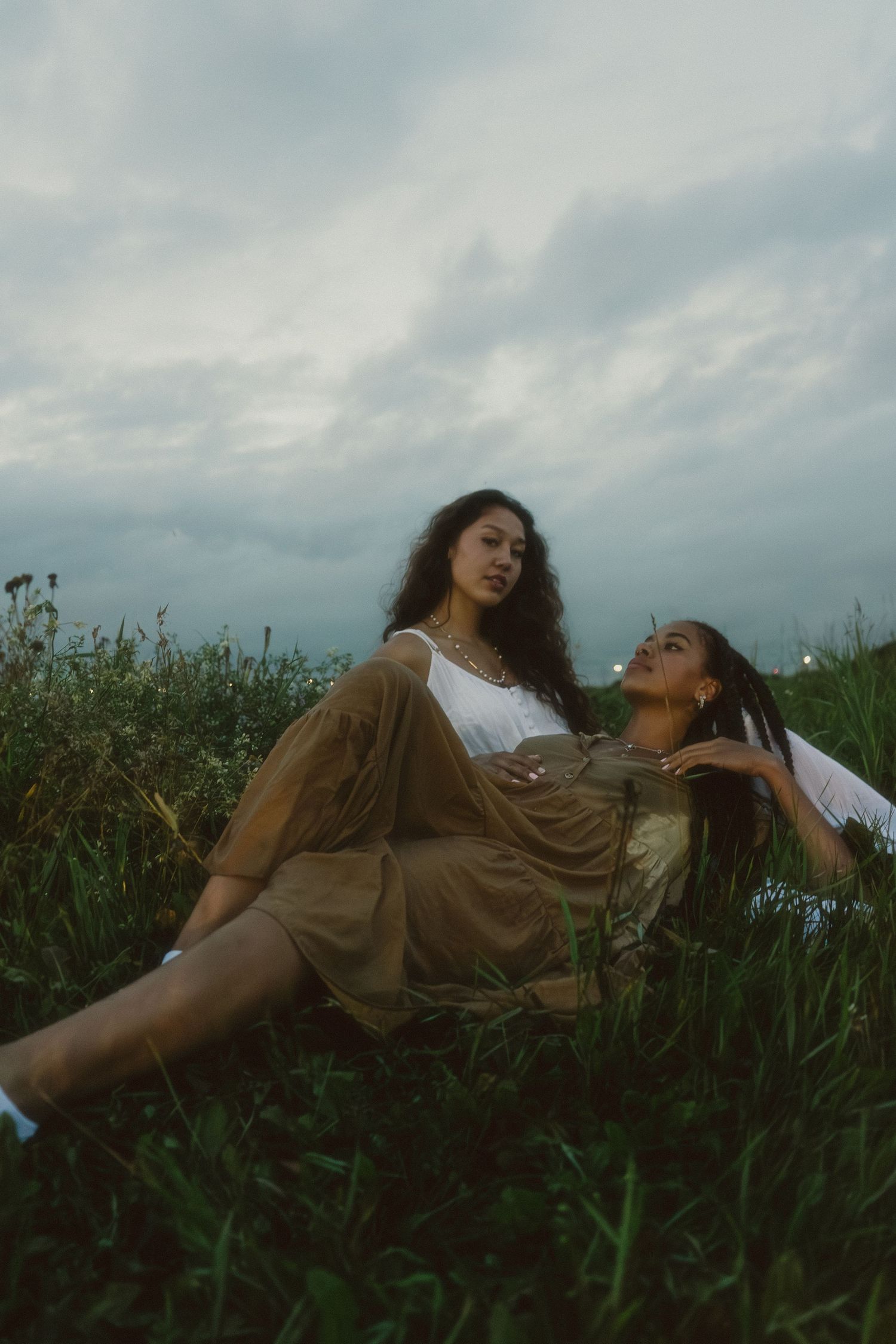 Two young women relax in a grassy field under a cloudy sky wearing earthy bohemian outfits.