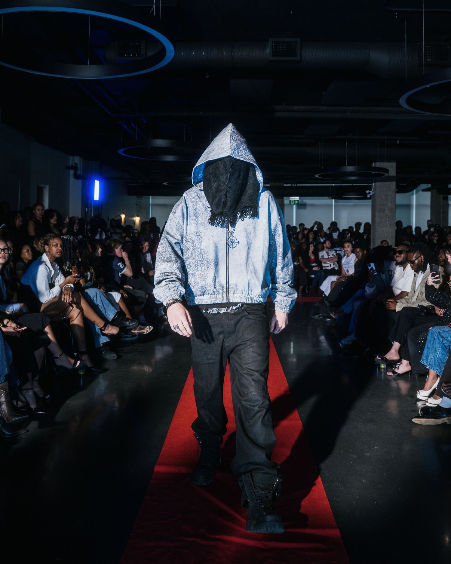 Model walks runway in silver reflective oversized hoodie with face covered and black pants at fashion show.