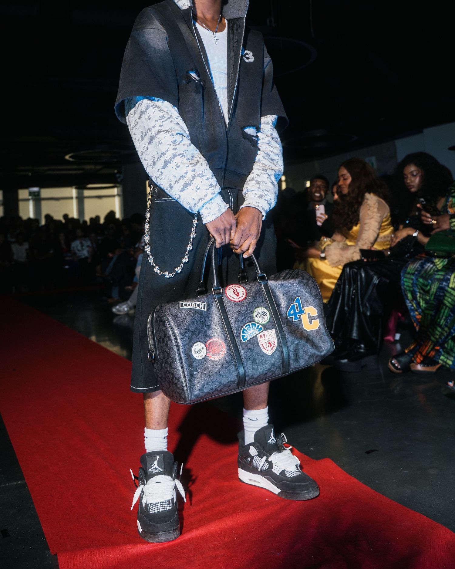 Close-up of model holding Coach duffle bag with patches, wearing Jordan 4 sneakers on red carpet runway.