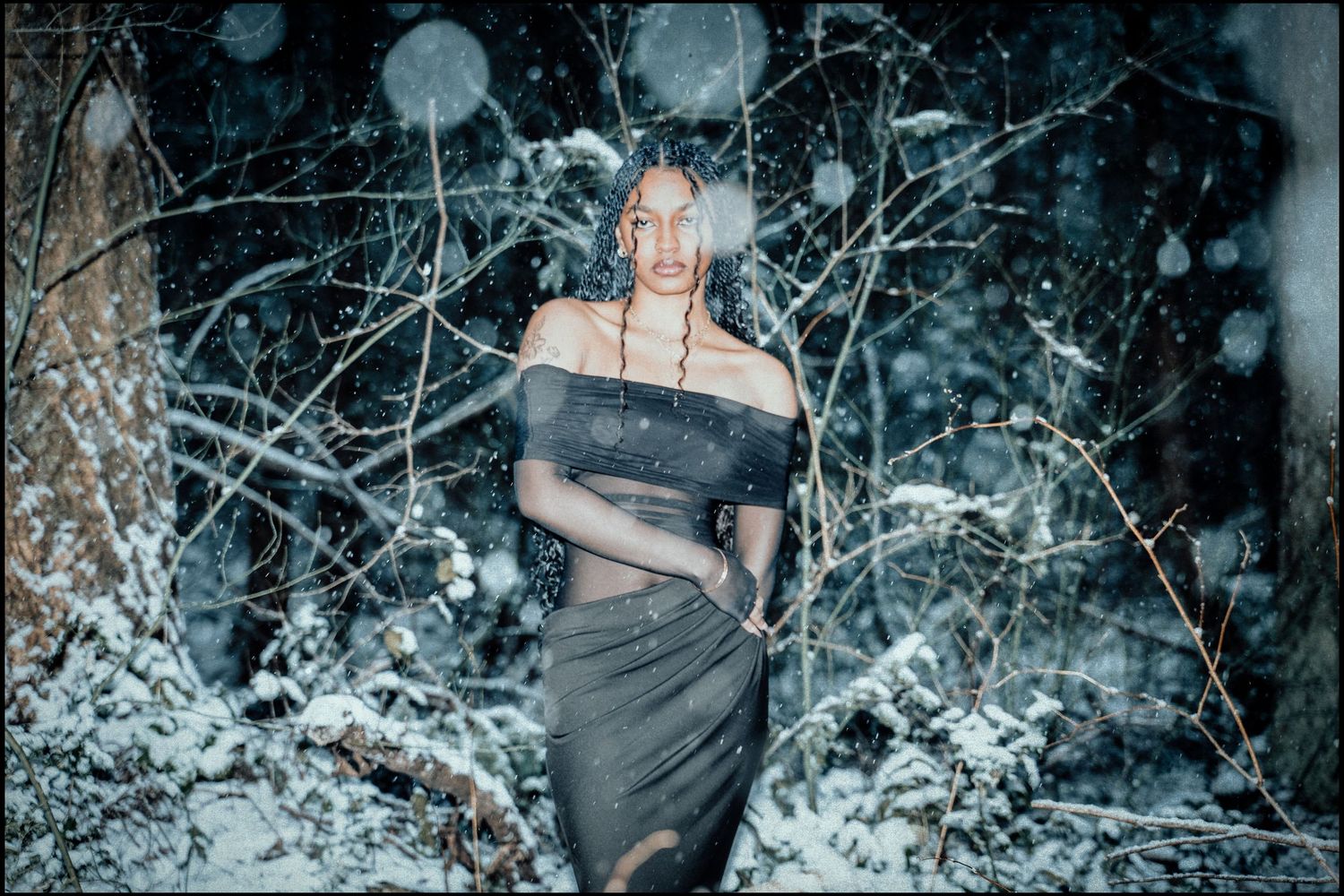 Woman in grey off-shoulder dress stands among snow-covered branches in winter forest.