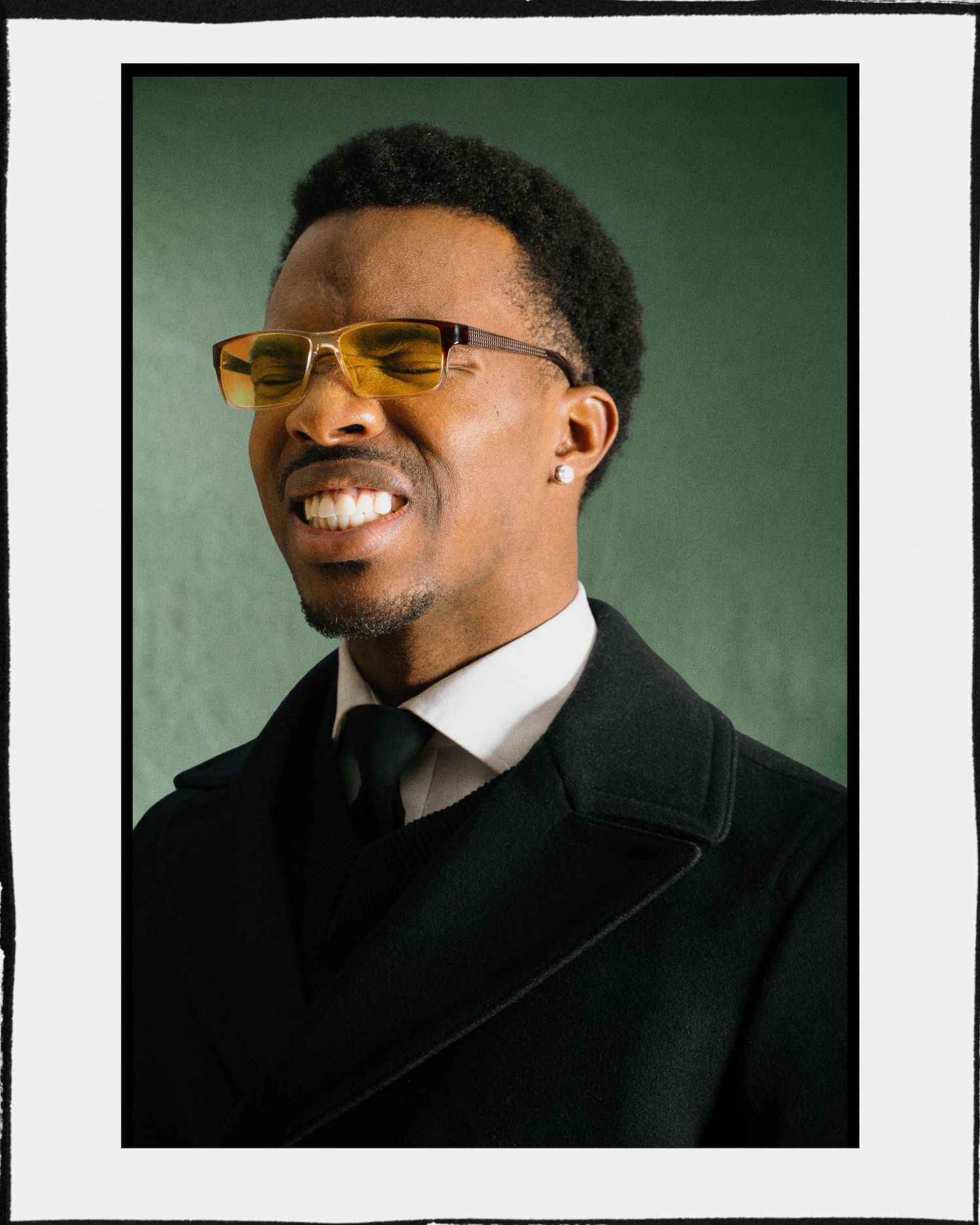 Smiling man wearing yellow tinted sunglasses and black coat against green background.