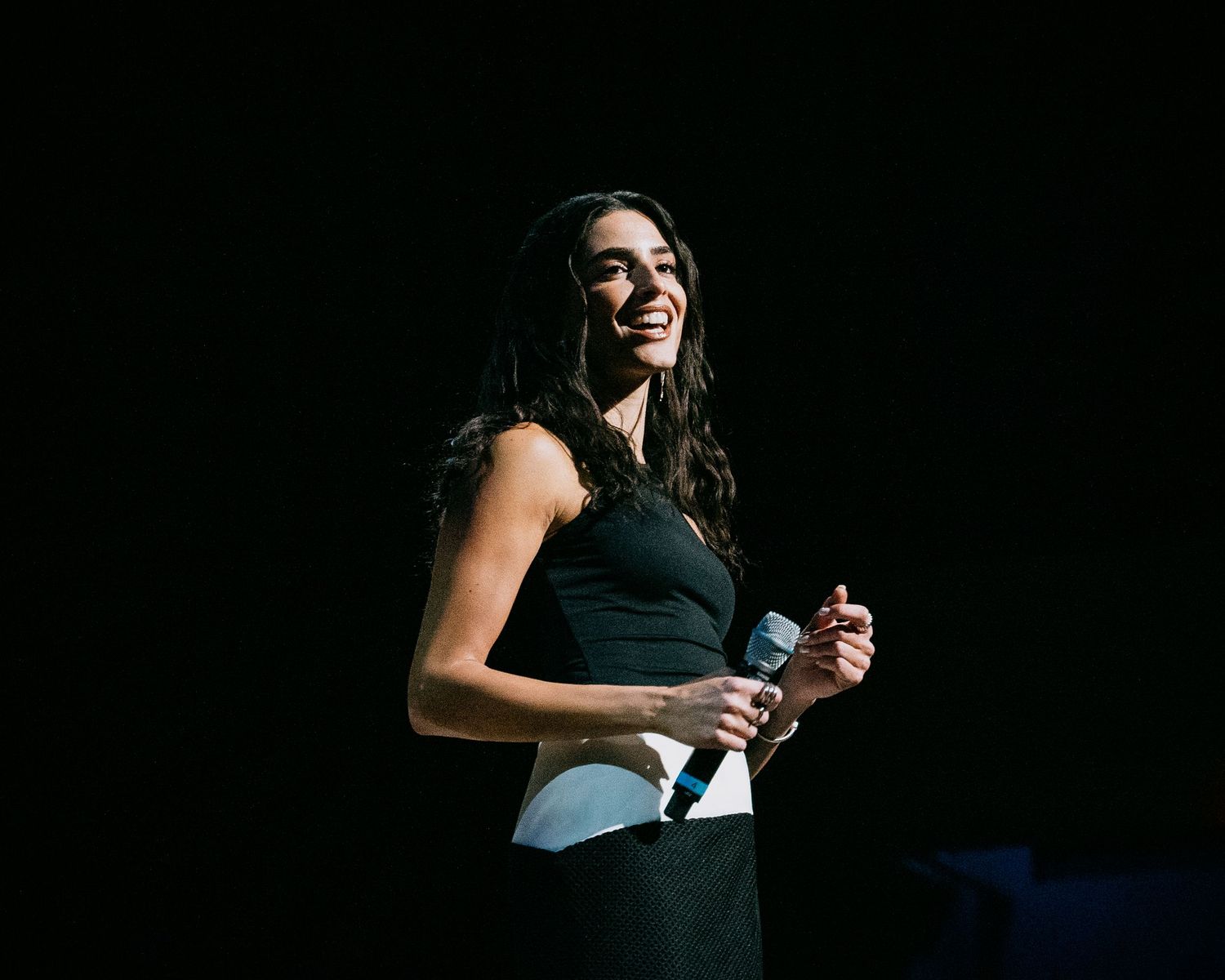 Smiling woman in black dress holds microphone on dark stage during a live performance or hosting event.