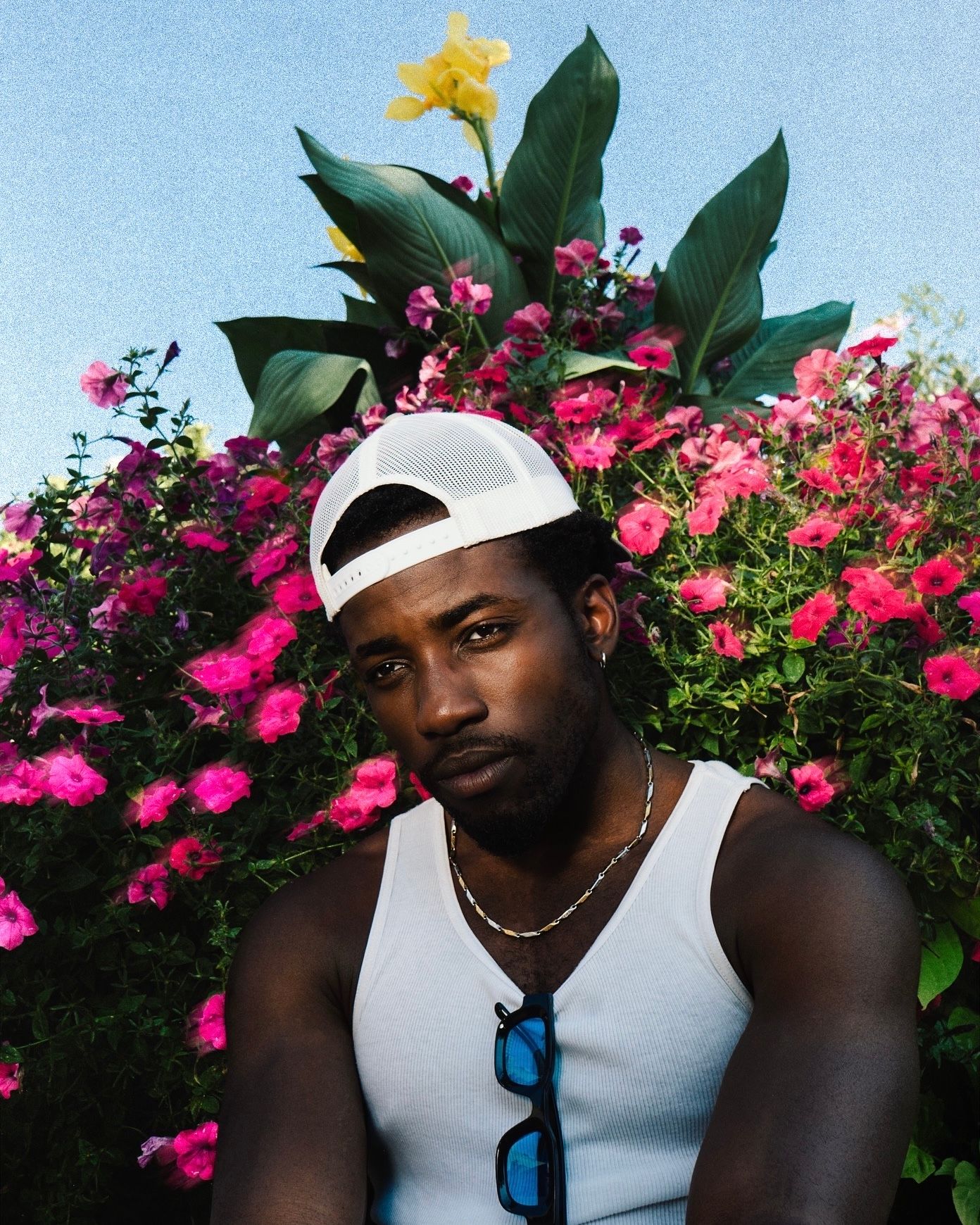 Man in white tank top and white cap posing in front of colorful pink and yellow flowers.