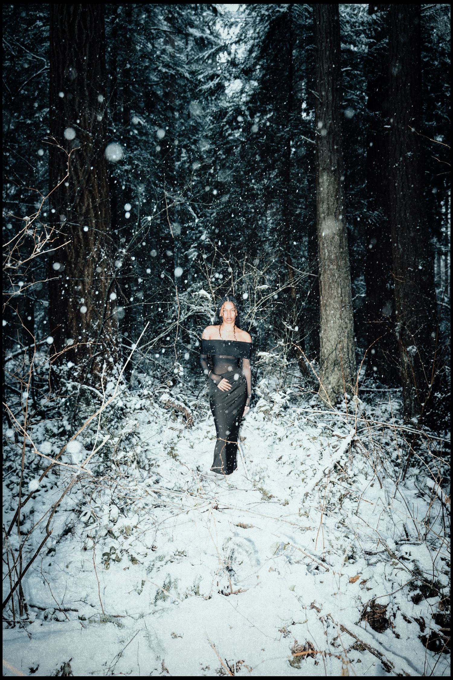 Woman in black outfit walking through snowy forest path with falling snowflakes.