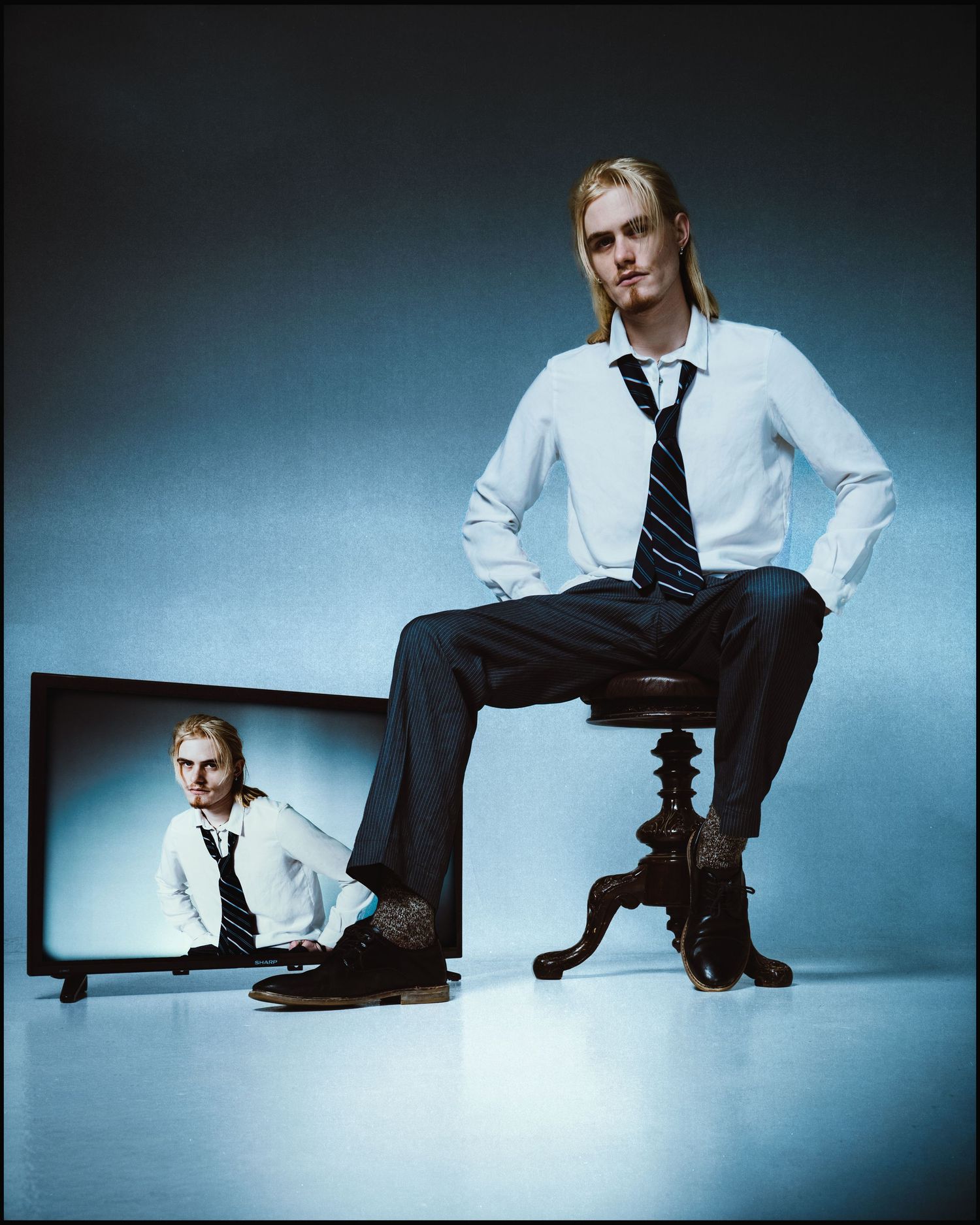 Blonde man in white shirt and tie sits on stool beside TV displaying his reflection in artistic portrait.