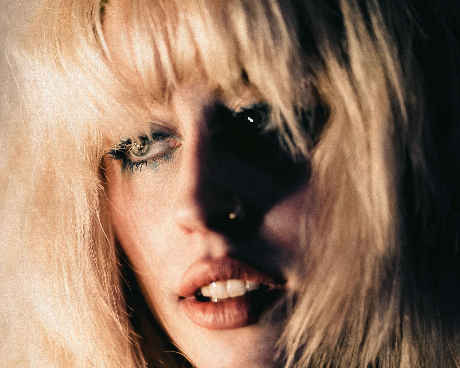 Close-up portrait of a blonde woman with dramatic eye makeup and a nose ring in moody lighting.
