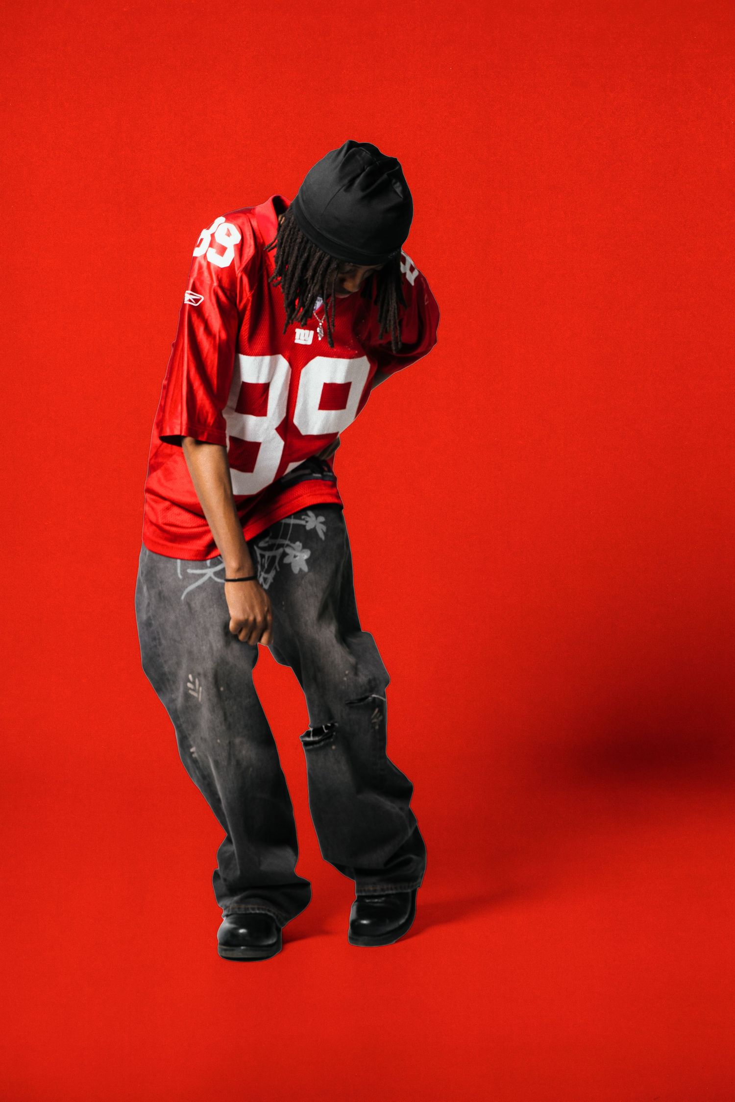 Person in red football jersey number 89, distressed jeans, and black beanie posing against red background.