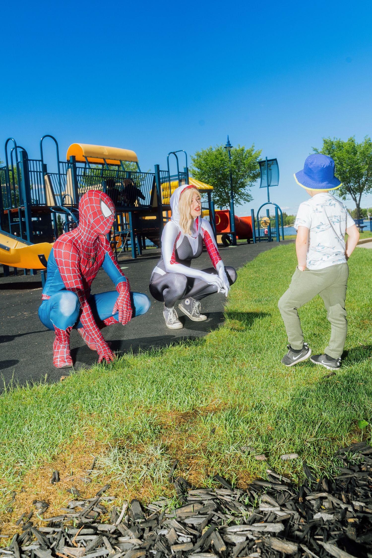 Two people in Spider-Man and Spider-Gwen costumes crouch to interact with a child at a playground.