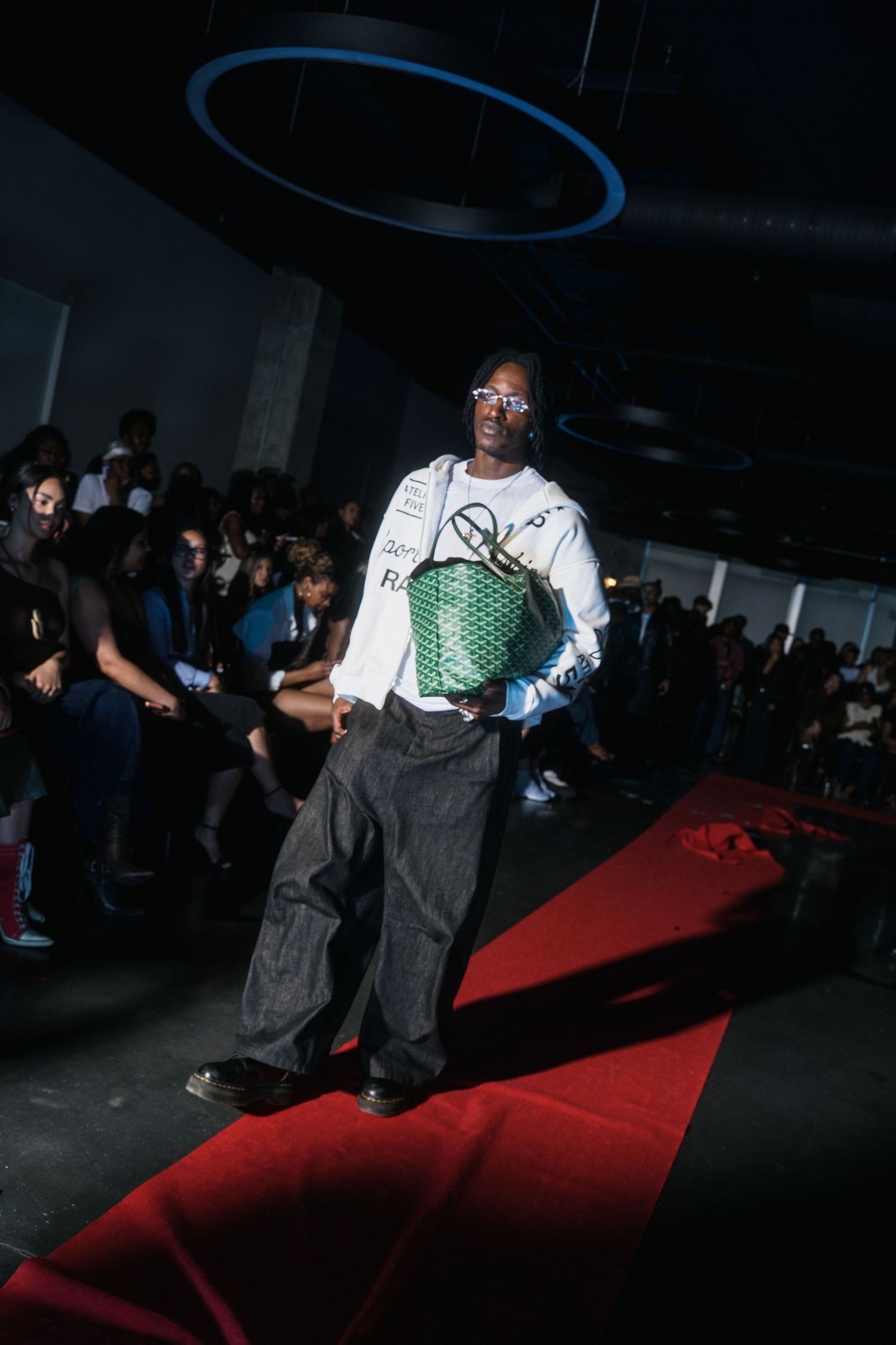 Male model in white graphic tee carrying a green Goyard bag on a red carpet fashion runway.