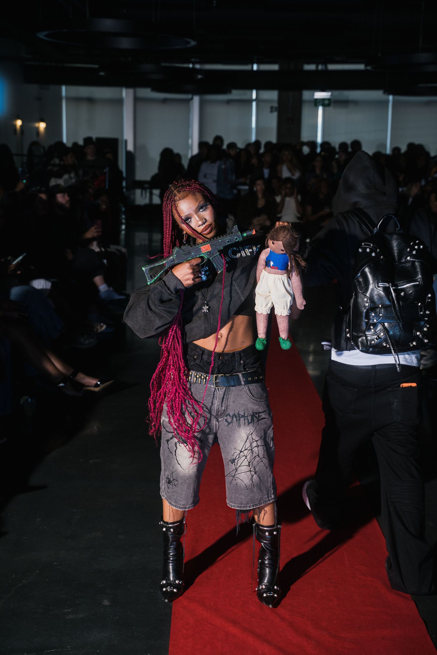 Female model with pink braids holding toy gun prop on red carpet runway at edgy fashion show.