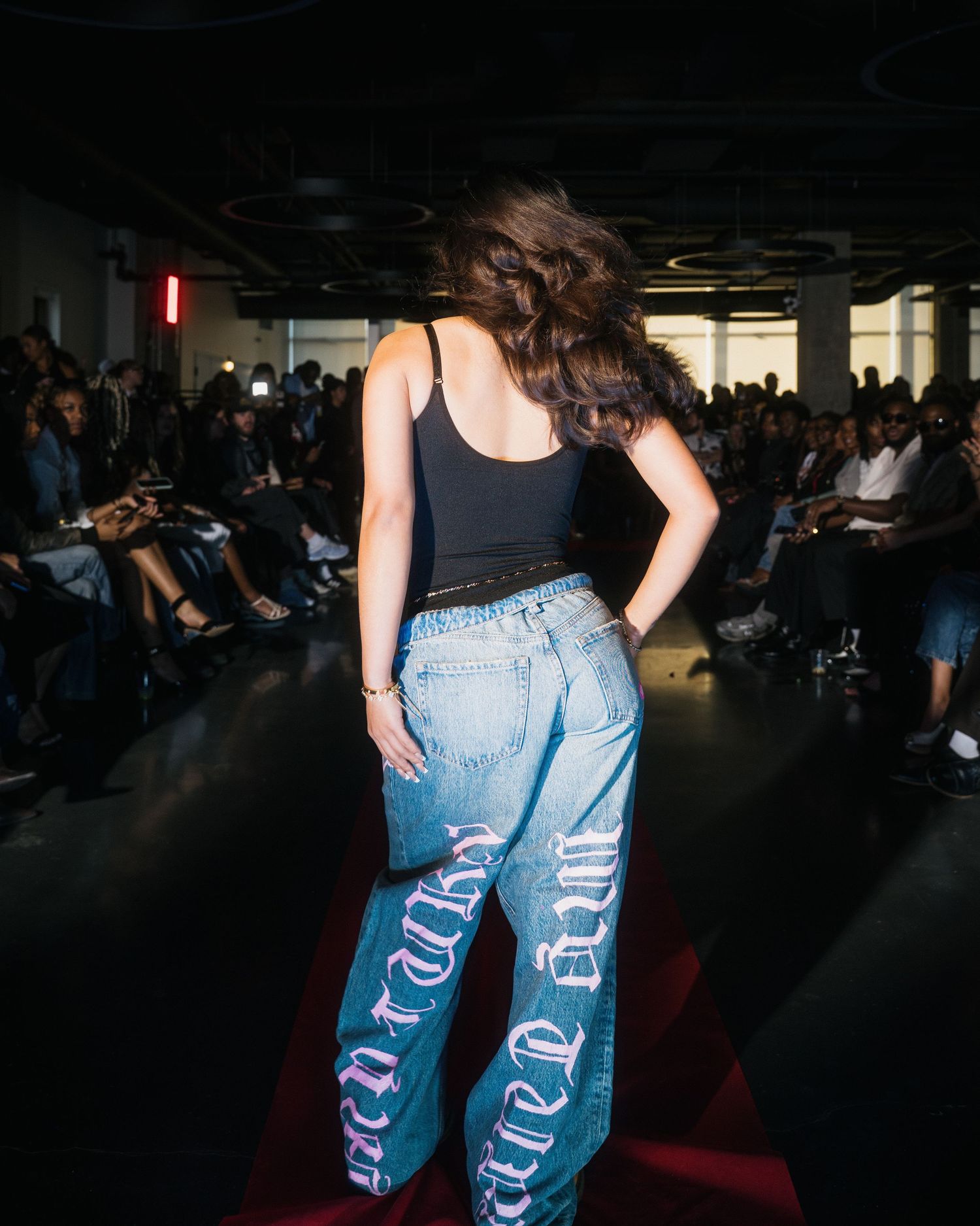 Model showcases baggy light wash jeans with gothic lettering graphic on fashion show runway.