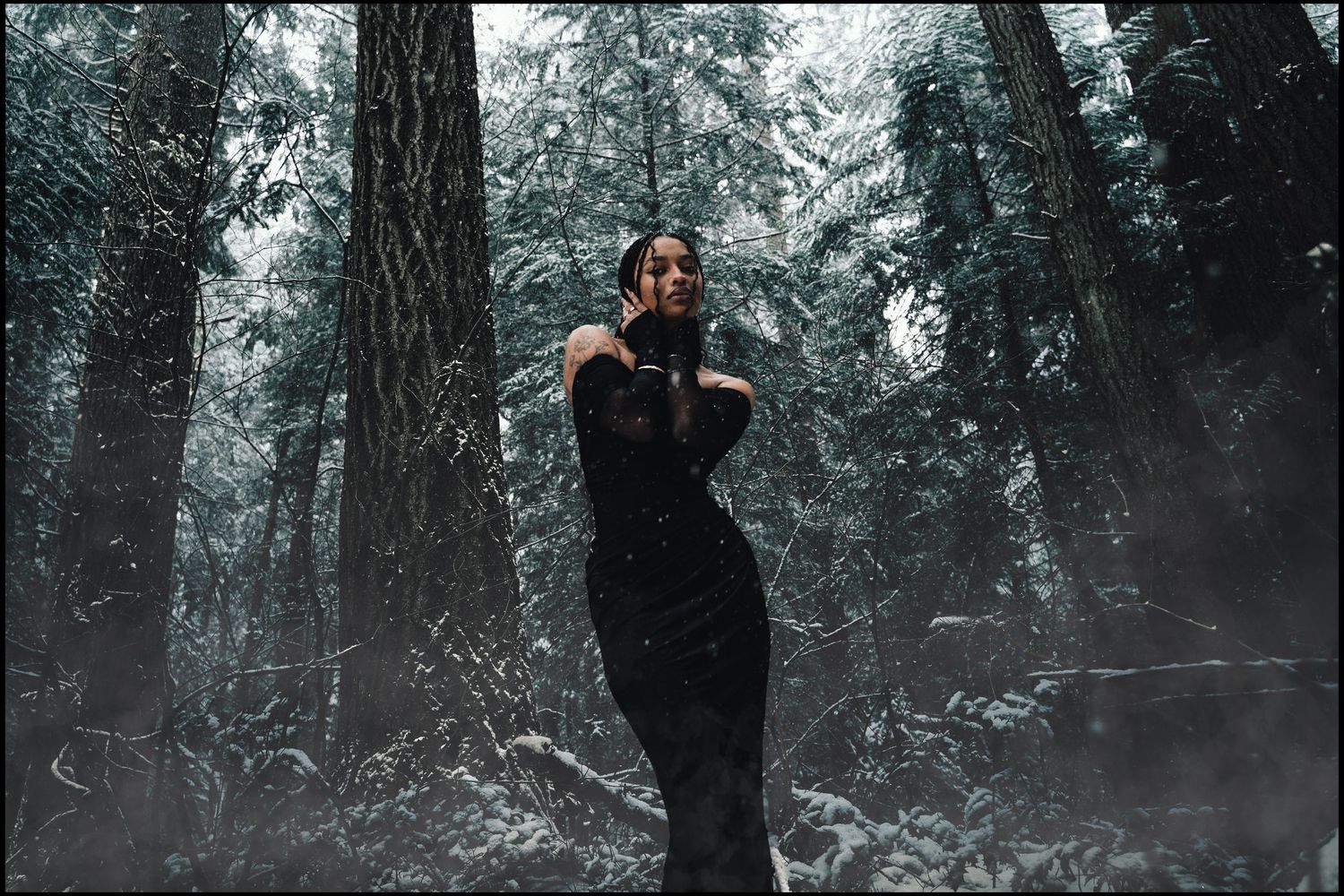 Woman in black dress posing dramatically in misty snowy forest setting.
