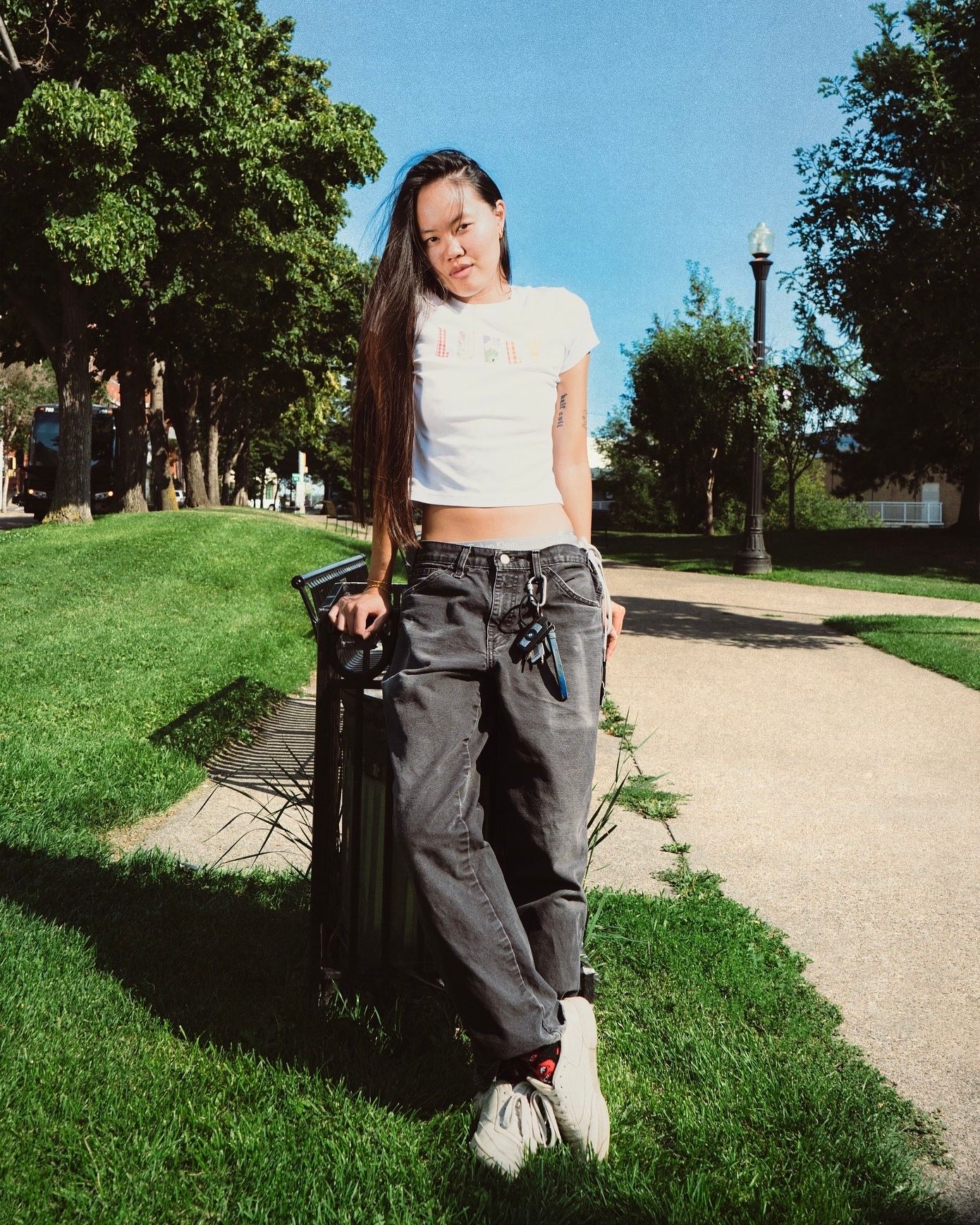 Young woman in white crop top and gray cargo pants standing on a park path on a sunny day.