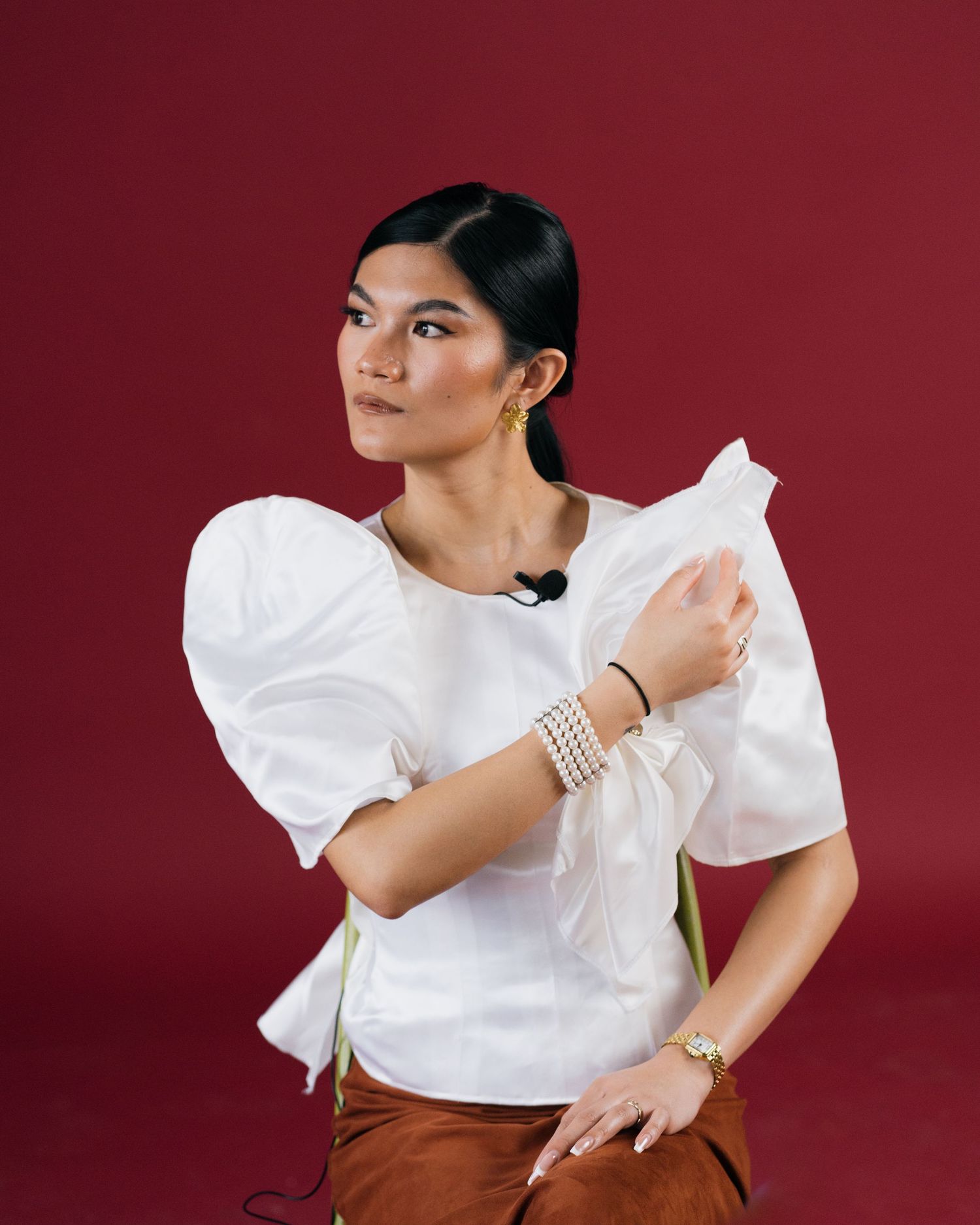 Woman in white puff-sleeve top with pearl bracelet and gold earrings on red background.