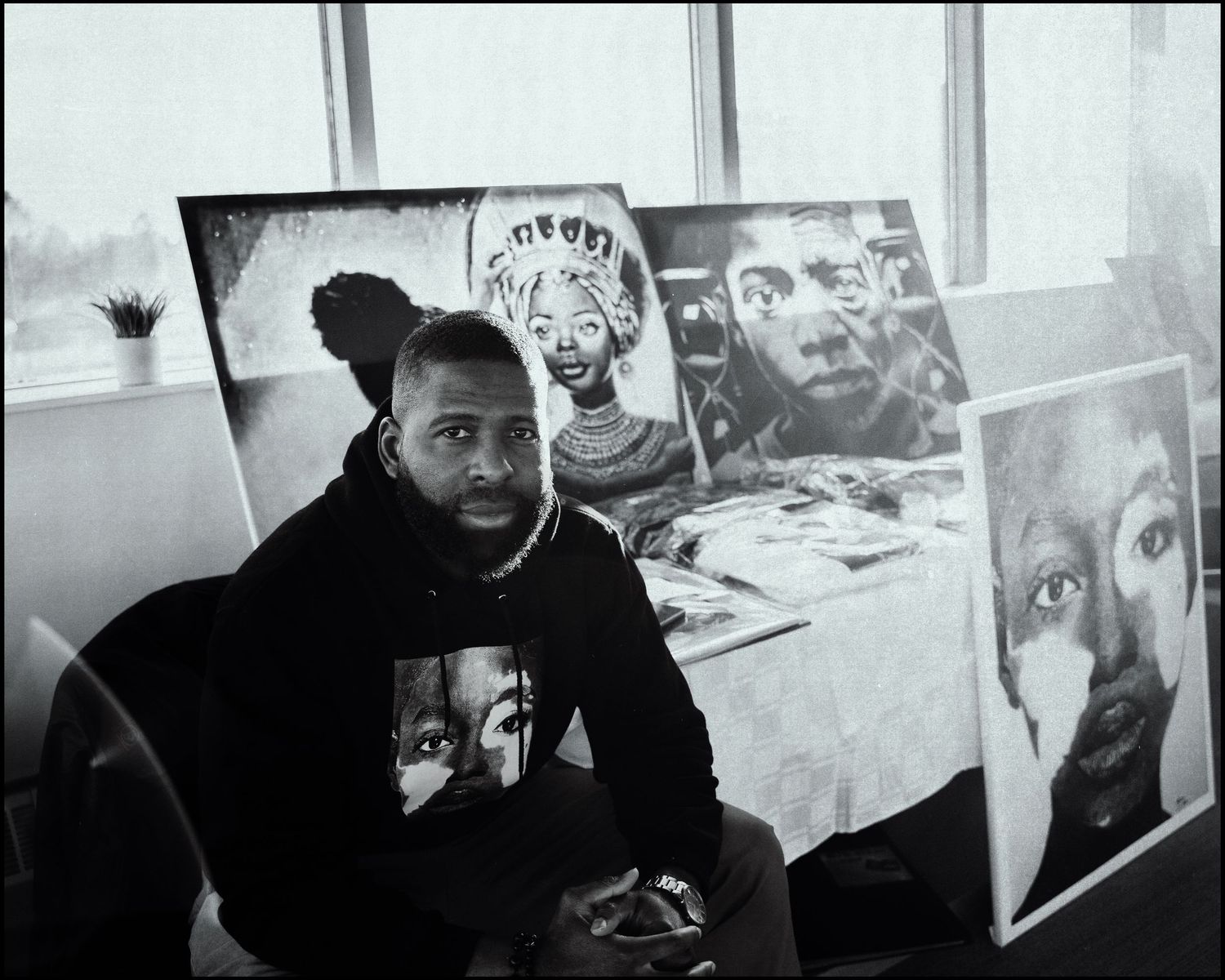 Black artist poses with large portrait paintings in monochrome studio setting.