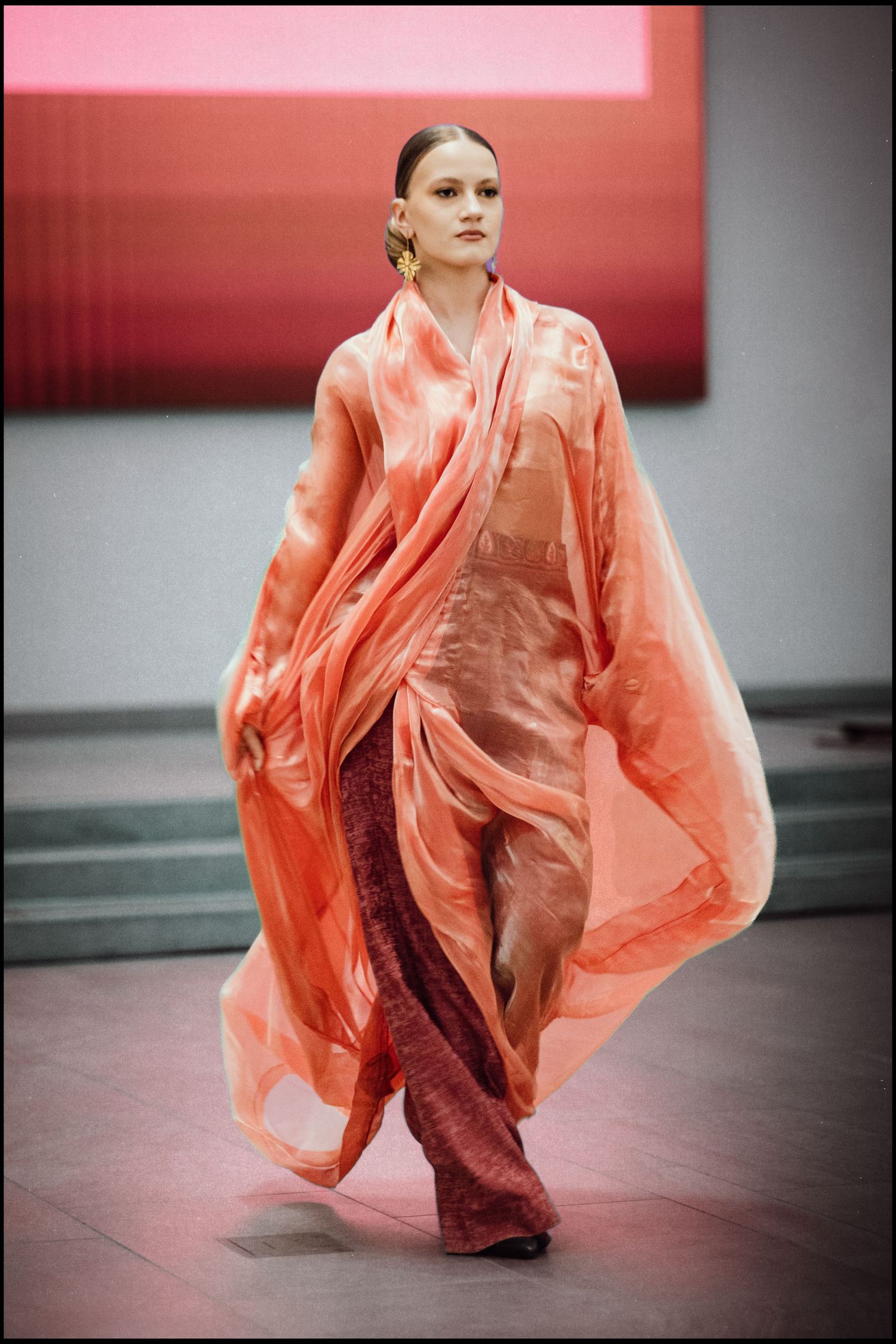 Model walks runway in flowing coral pink sheer organza kimono-style gown with wide sleeves over burgundy trousers.