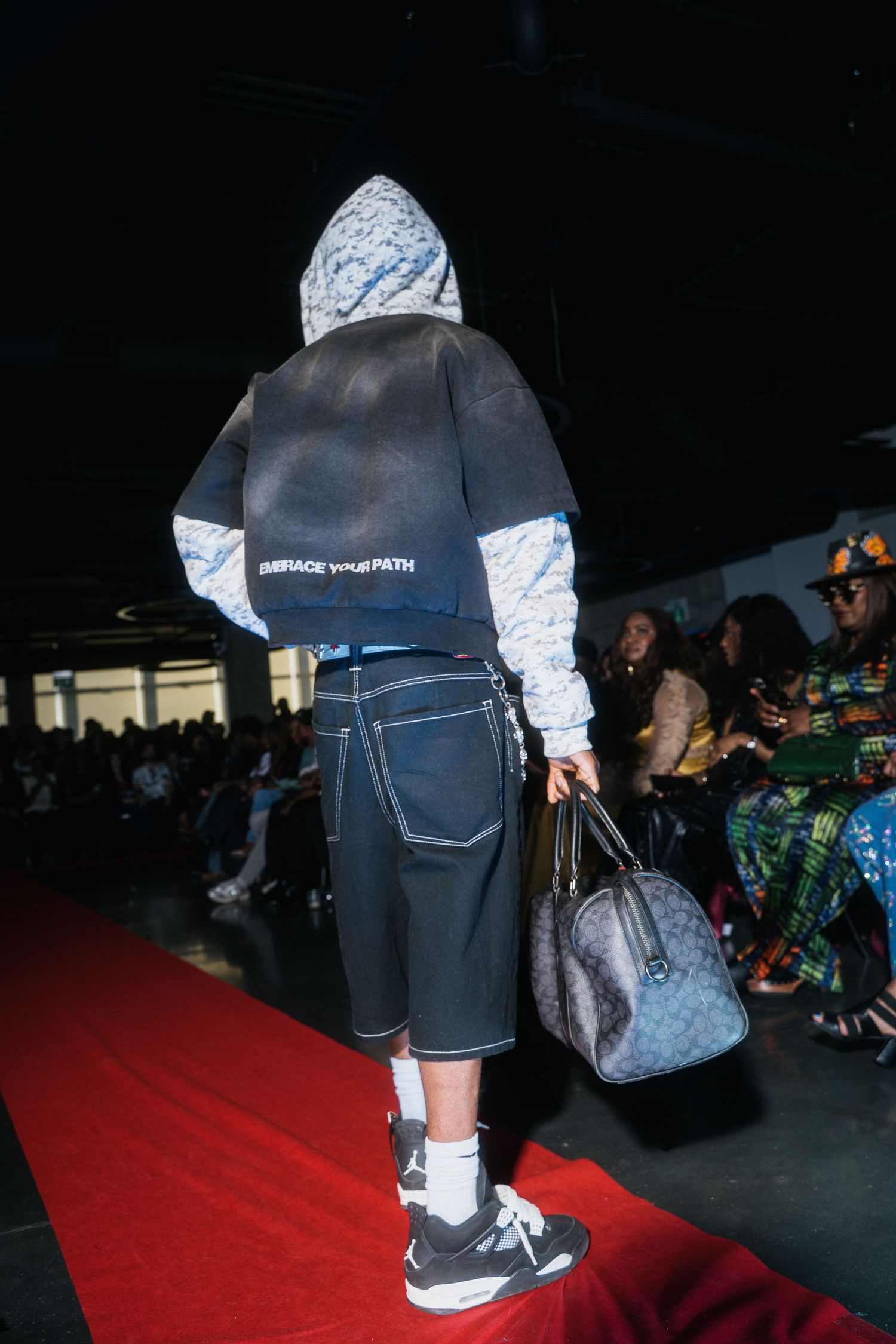Back view of model in two-tone hoodie reading Embrace Your Path, carrying duffle bag on runway.