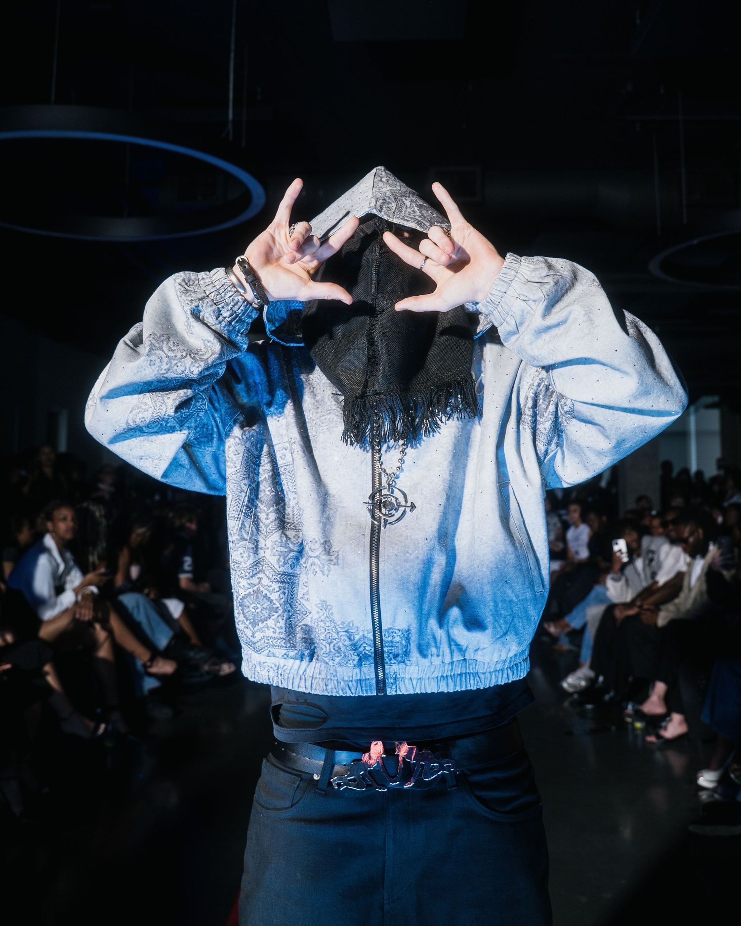 Model poses on runway covering face with hands wearing silver paisley bomber jacket at fashion show.