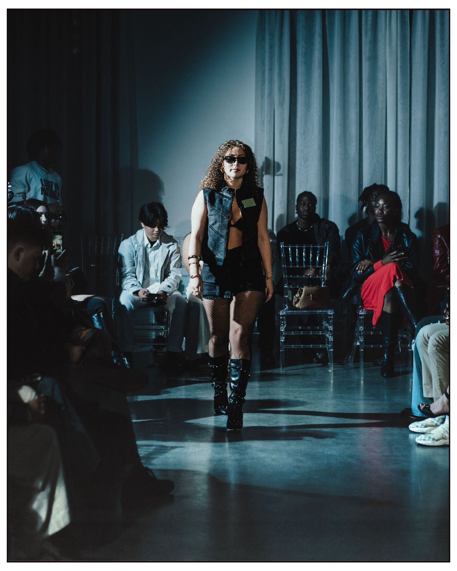 Model struts runway in black denim vest and shorts with knee-high boots at fashion show.