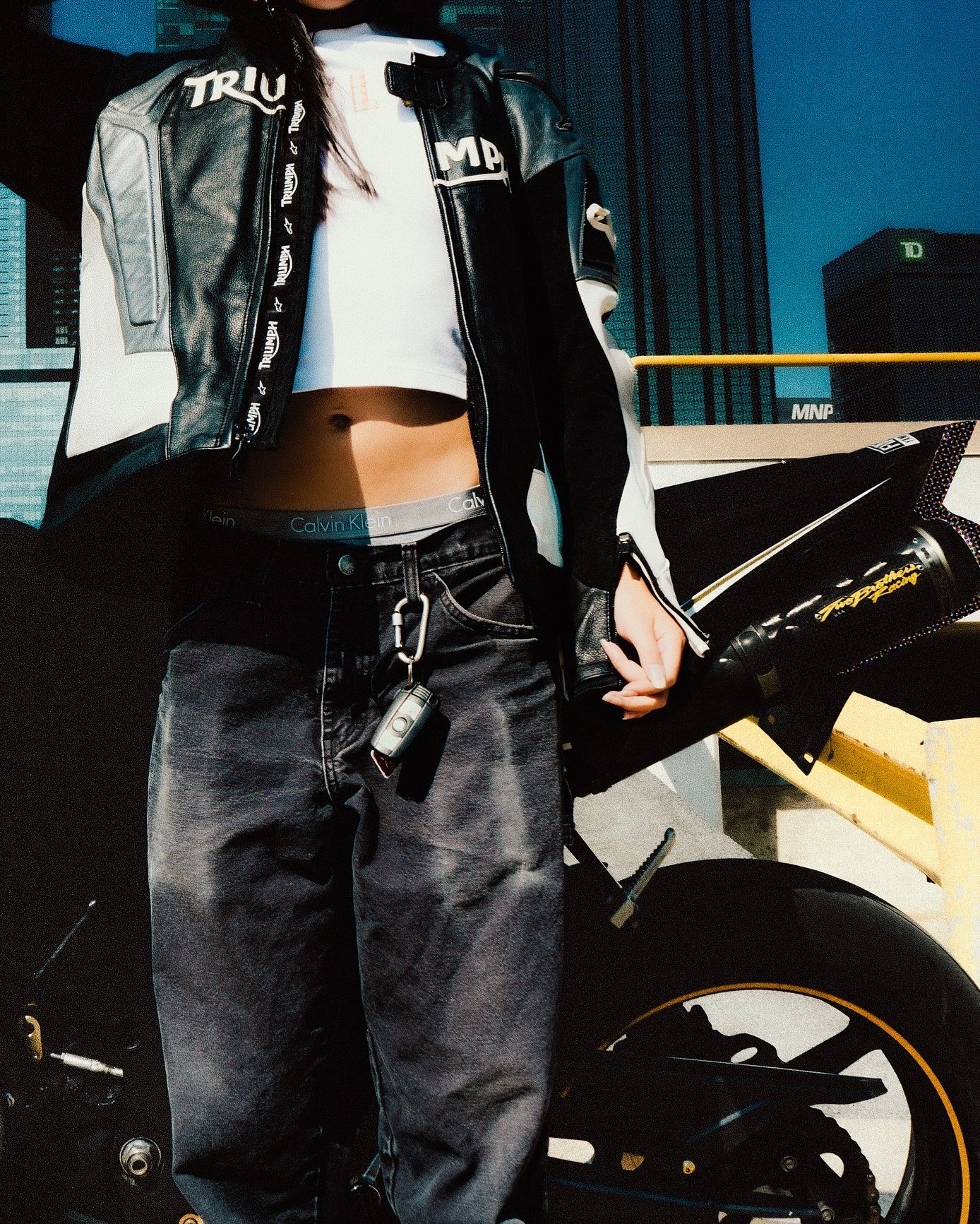 Woman in black leather jacket and gray jeans posing beside a motorcycle in an urban setting.