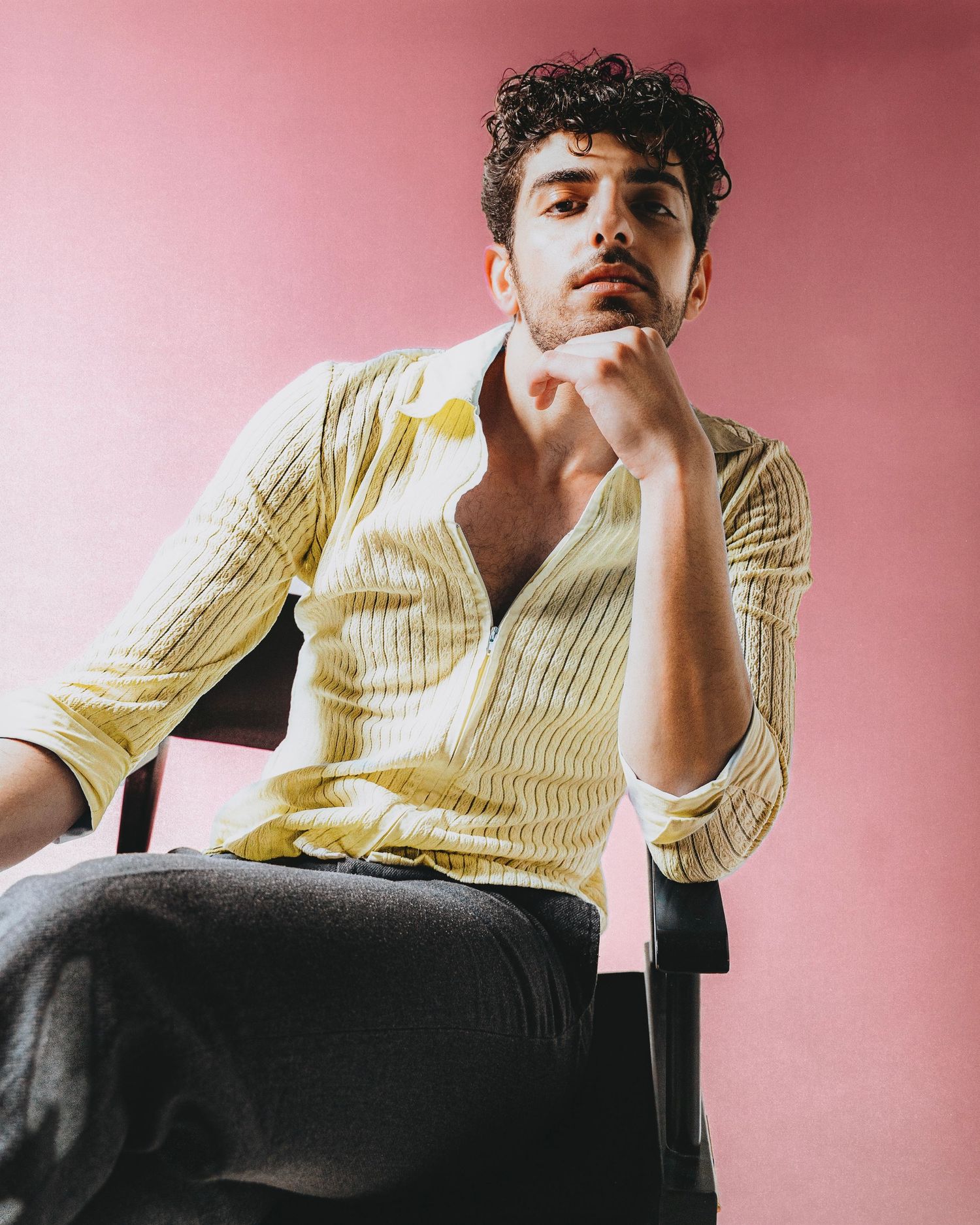 Young man with curly hair in yellow striped shirt sits on chair against pink background.