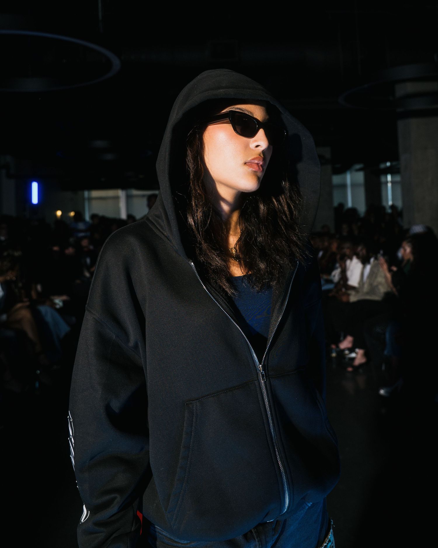 Model poses in black oversized zip hoodie and sunglasses at urban streetwear fashion runway show.