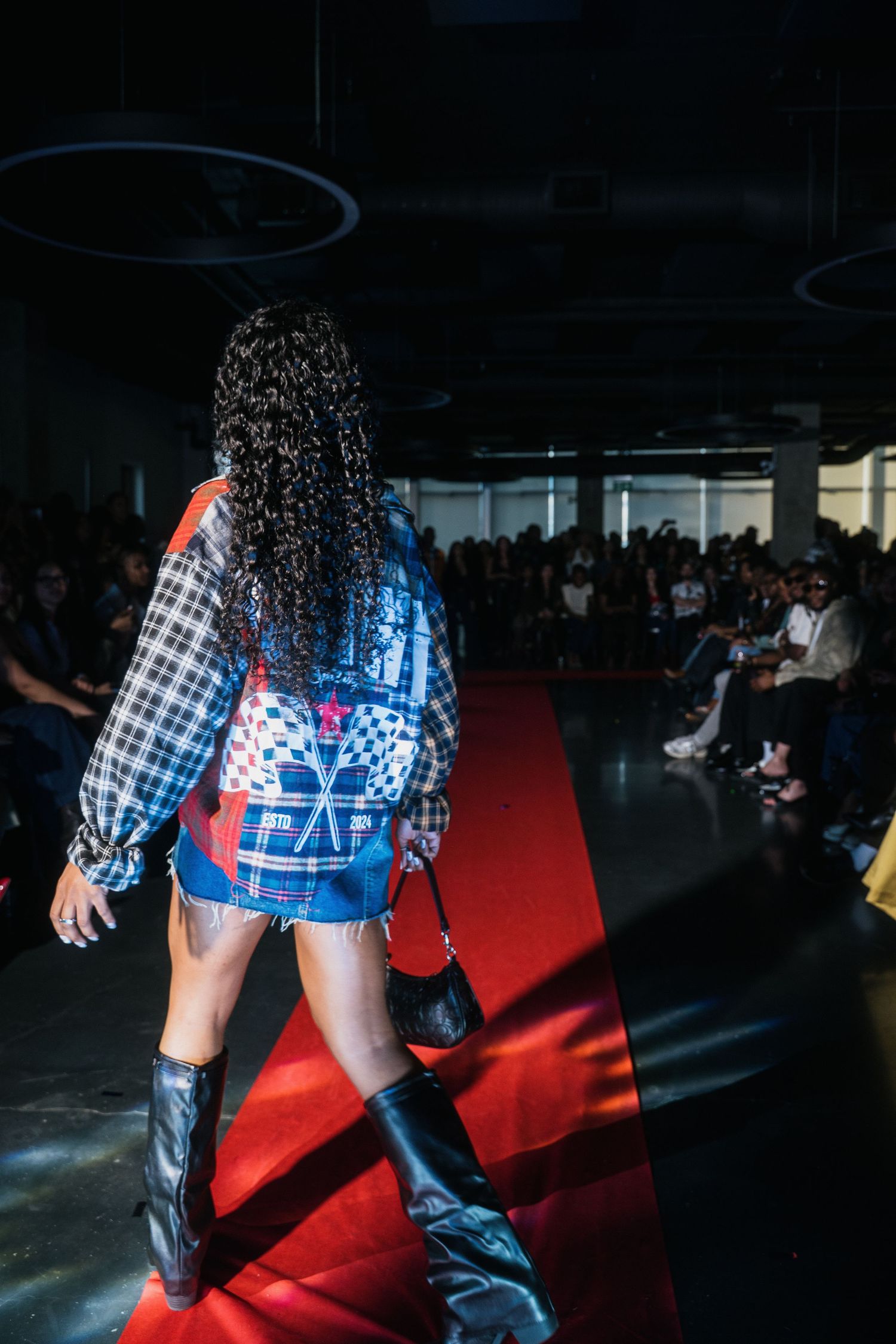 Model walks red carpet runway in patchwork plaid jacket, denim shorts, and black knee-high boots.