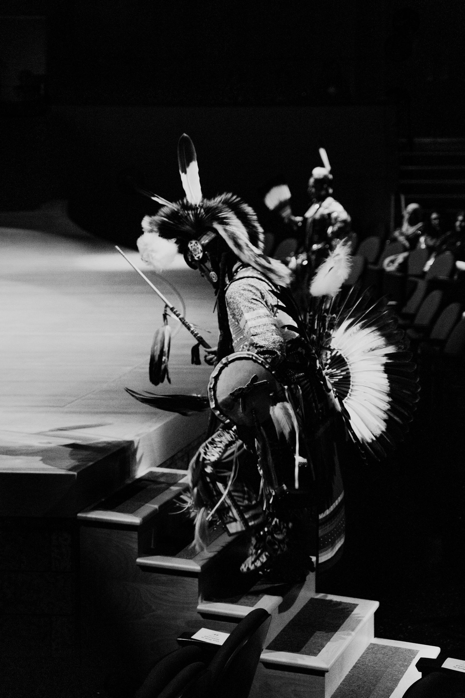 Black and white photo of Indigenous dancer in traditional regalia with feathers performing at a powwow ceremony.