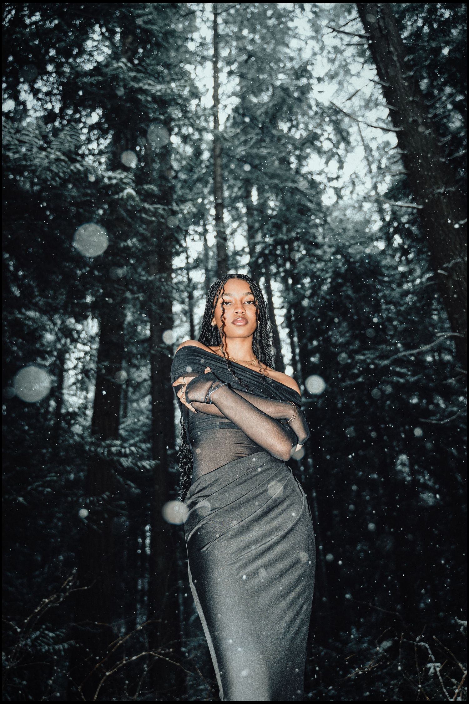 Woman in grey dress stands confidently among tall snow-covered trees in winter forest.