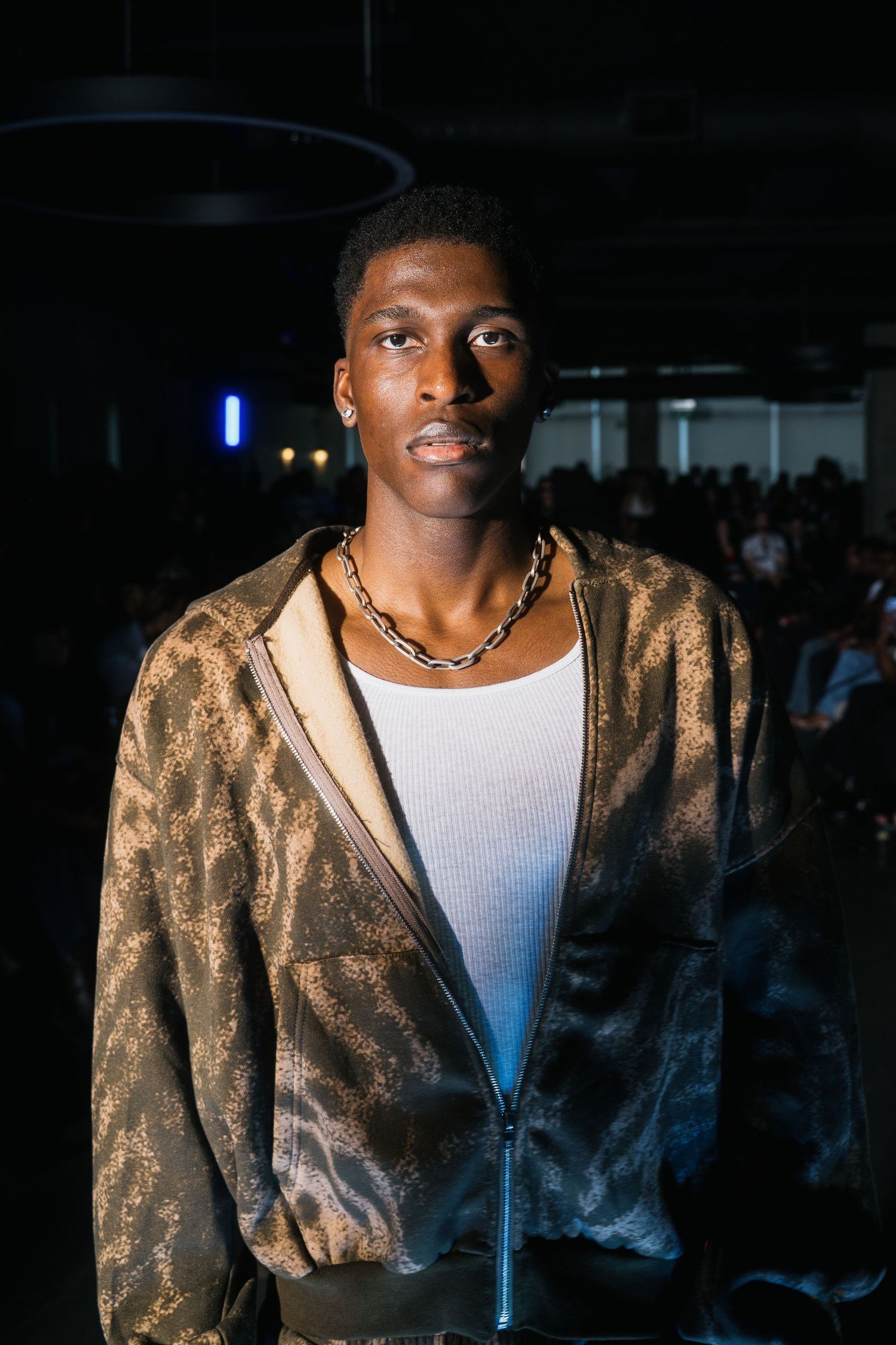 Male model walks runway in brown leopard print bomber jacket with chain necklace at fashion show.
