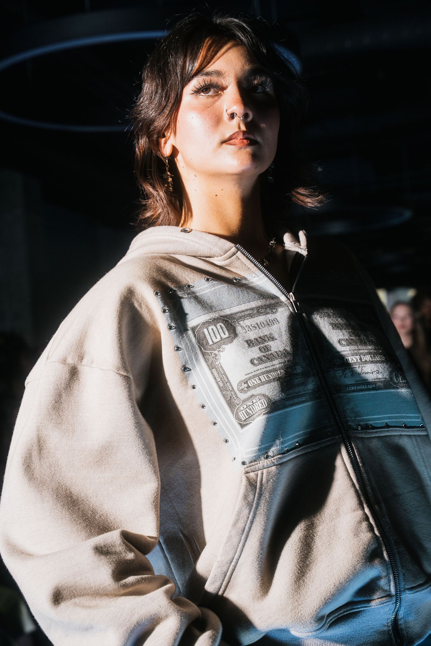 Model wears oversized beige hoodie with $100 bill graphic print at fashion show.