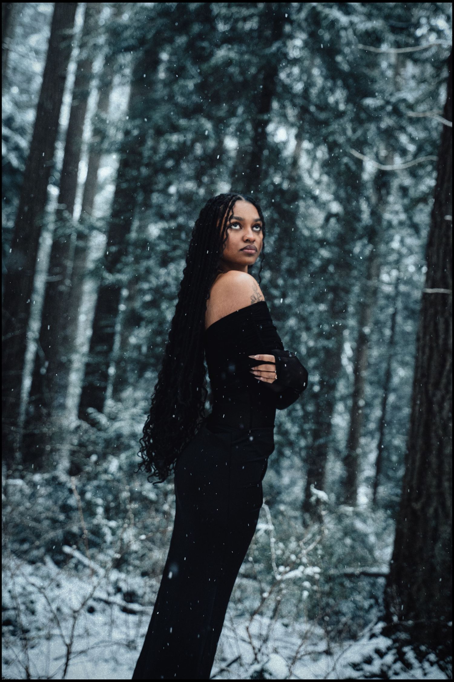Woman in black dress with long braids looking back in snowy forest.