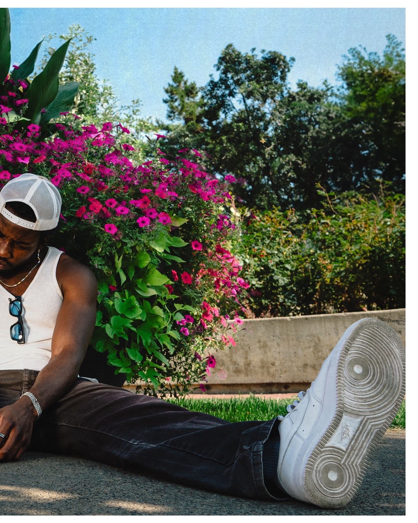 Man in white tank top and white sneakers relaxing by vibrant pink flowers in a garden.