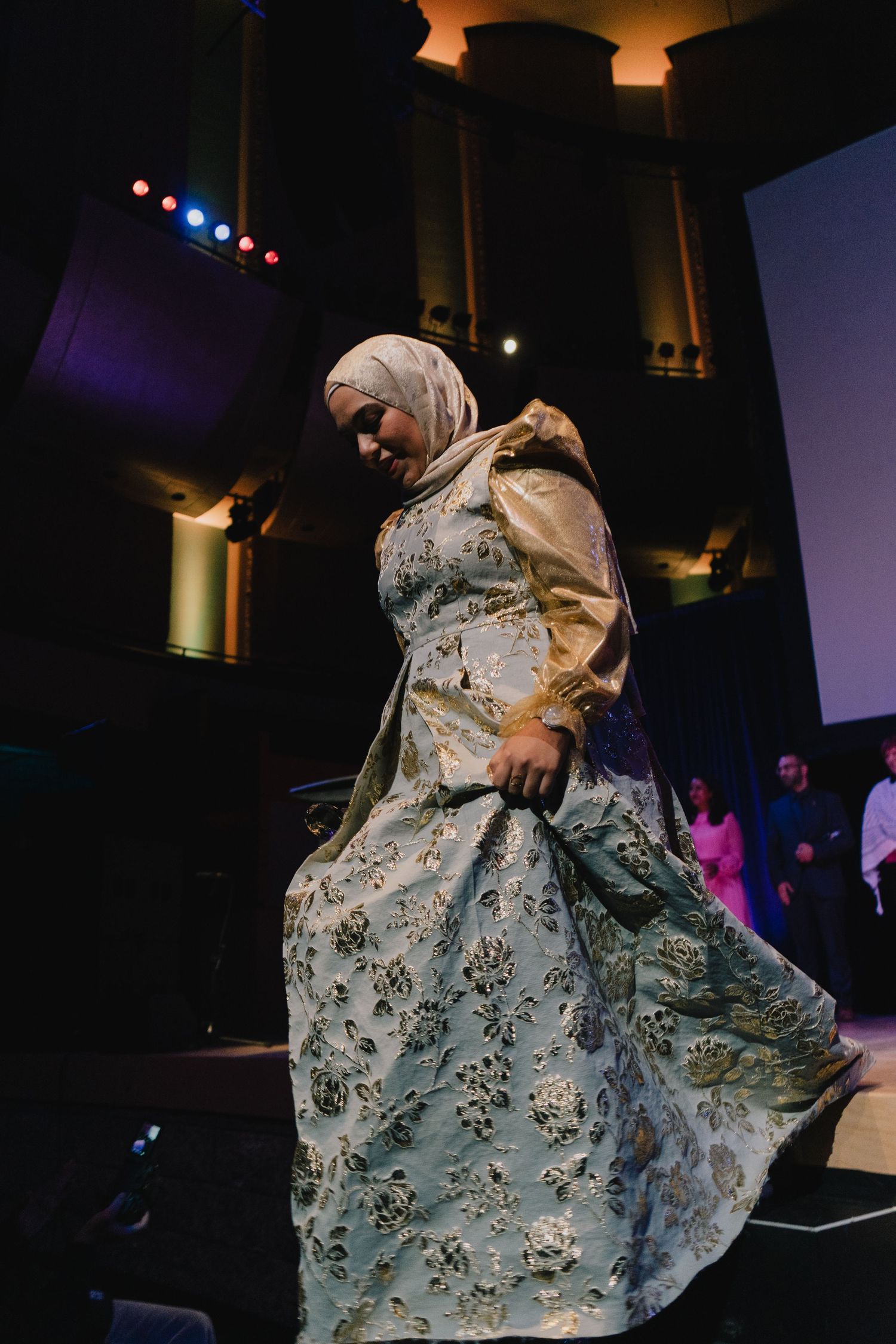 A woman in a hijab and elegant floral gown twirls gracefully on a stage under colorful lights.