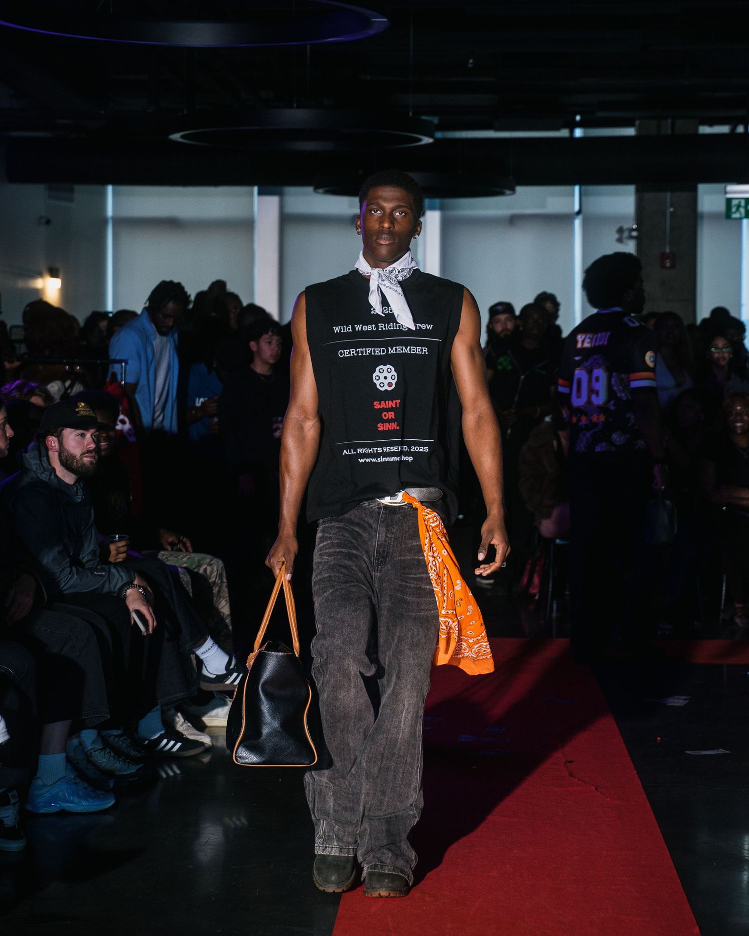 Male model walks red carpet runway in graphic tank top, wide-leg pants, and carrying black tote bag.